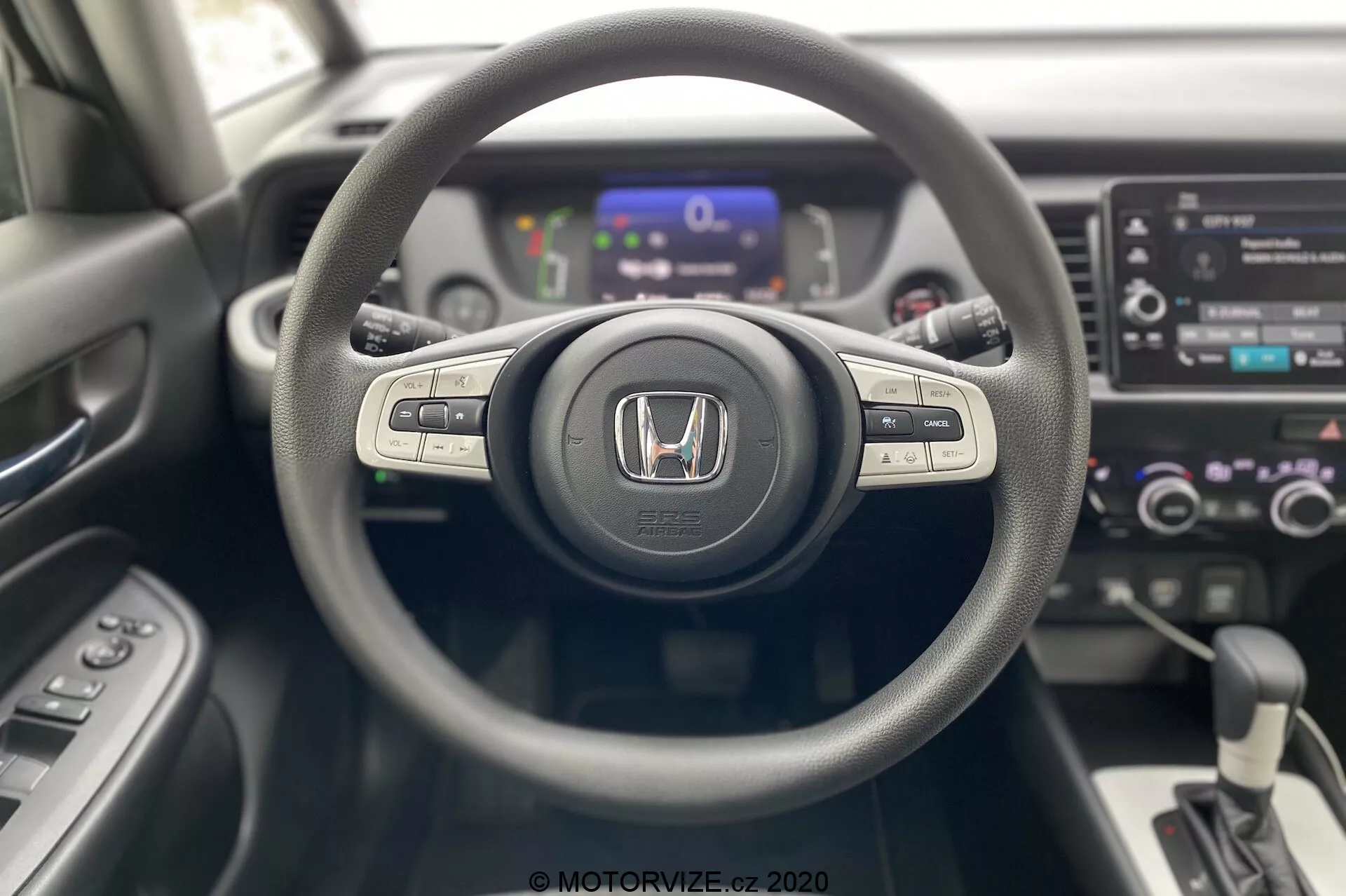 Vue intérieure de la Honda Jazz 2020 depuis la perspective du conducteur, mettant en avant le volant à trois branches avec emblème Honda, les boutons de commande du système multimédia et du régulateur de vitesse sur les branches, un tableau de bord numérique légèrement flou en arrière-plan et une console centrale avec système d'info-divertissement à écran tactile, commandes de climatisation bi-zone et levier de vitesses automatique. Design moderne et contemporain avec des matériaux de textures mixtes.