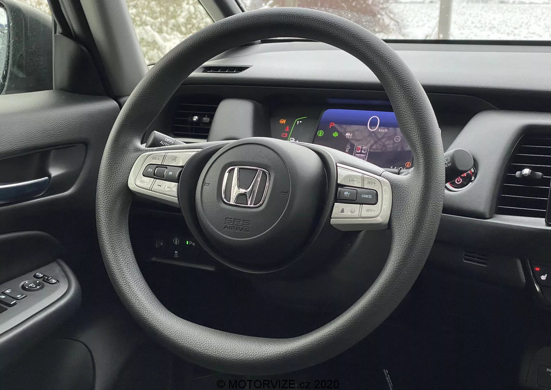 Vue intérieure d'une Honda Jazz 2020 depuis la perspective du siège du conducteur, mettant en avant le volant à trois branches avec le badge Honda et des commandes intégrées pour l'audio, le régulateur de vitesse et probablement le Bluetooth ou la commande vocale. Derrière le volant se trouve un tableau de bord numérique avec des affichages colorés incluant un compteur de vitesse et un compte-tours. Partiellement visible, la console centrale abrite un écran d'infodivertissement ou de navigation et des bouches de contrôle de la climatisation. Le panneau de la porte du conducteur avec boutons de contrôle des fenêtres et des rétroviseurs latéraux est également partiellement visible.
