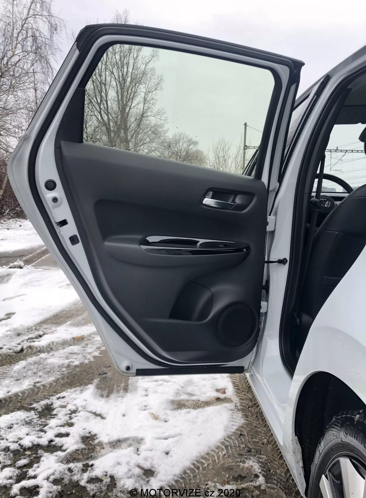 Gros plan sur l'intérieur de la porte avant côté conducteur de la Honda Jazz 2020, montrant une vue détaillée de la finition en plastique texturé, de la poignée ergonomique satinée, des commandes de fenêtre et de rétroviseur électriques, de la poche de rangement moulée et de la grille de haut-parleur, le tout dans un décor hivernal enneigé.
