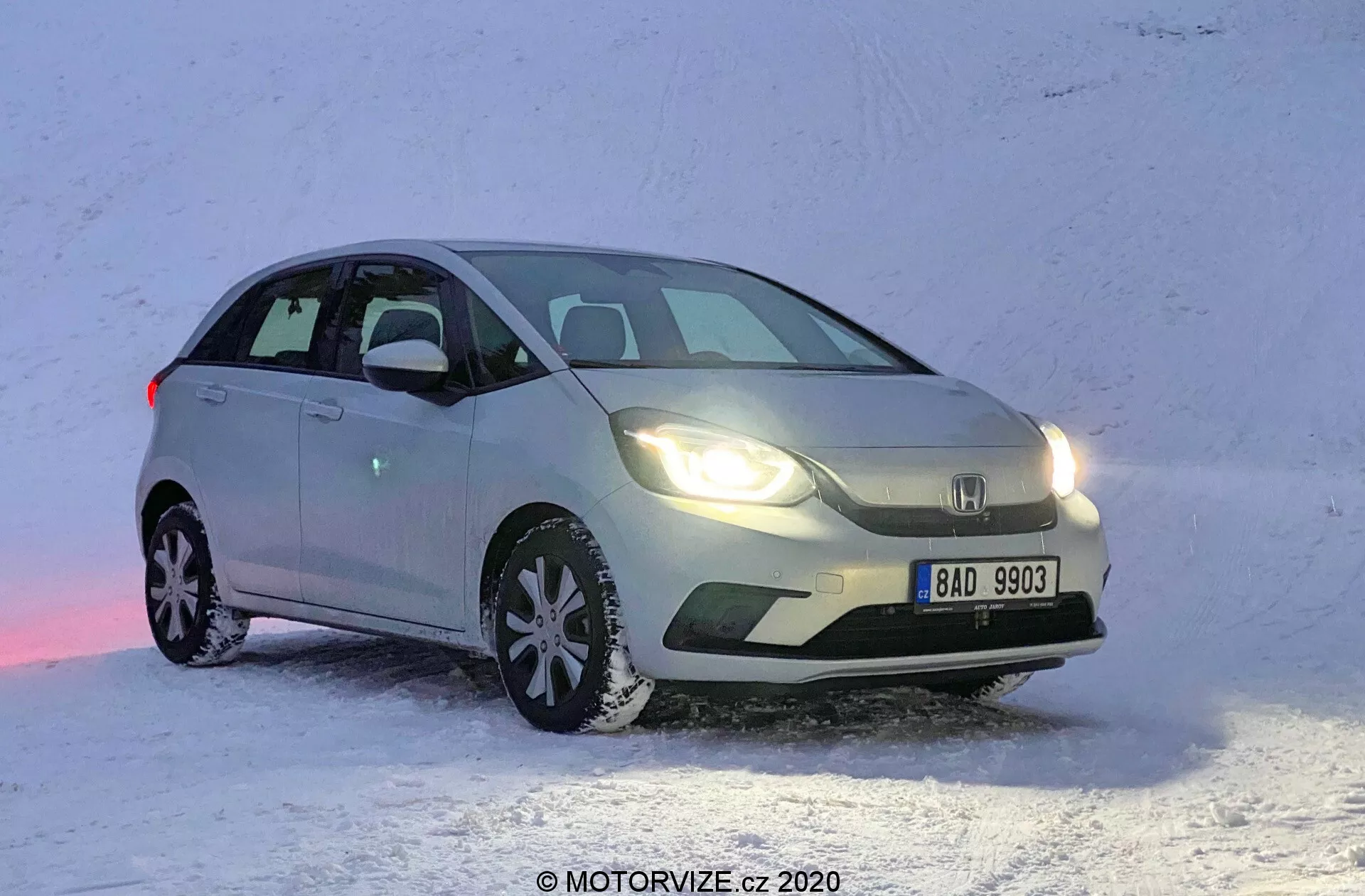 Vue de trois quarts frontale d'une Honda Jazz 2020 de couleur claire, positionnée sur une surface enneigée. L'image capture la voiture du côté avant gauche, montrant le côté conducteur sous un angle qui met en valeur à la fois le profil latéral et la façade avant. La Honda Jazz est garée avec le front légèrement tourné vers la droite, révélant le design de la grille, des phares et d'une partie du capot. Les phares avant sont allumés, reflétant sur la neige, avec le véhicule apparemment à l'arrêt. Les pneus neige avec des motifs de bande de roulement distinctifs sont visibles, soulignant son aptitude à la conduite hivernale.