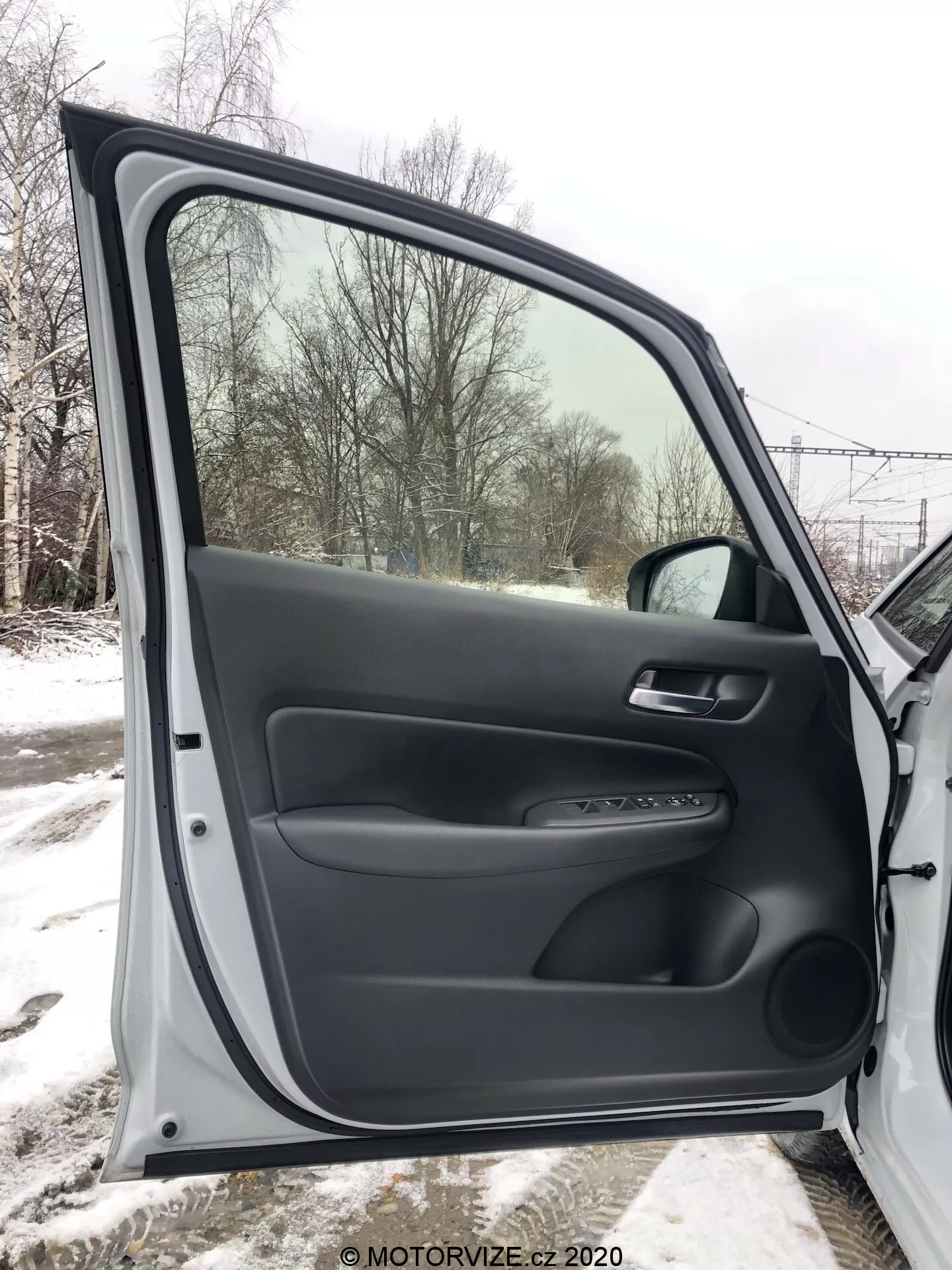 Vue intérieure de la porte avant passager du Honda Jazz 2020, entièrement ouverte, présentant des matériaux plastiques noirs texturés et lisses. Le panneau de porte comprend un accoudoir ergonomique, une poignée en matériau souple et un panneau de contrôle pour les mécanismes de fenêtre et de verrouillage électriques. Une garniture argentée et une poche de rangement inférieure sont également visibles. À travers la fenêtre, un paysage enneigé est visible, indiquant une ambiance hivernale.