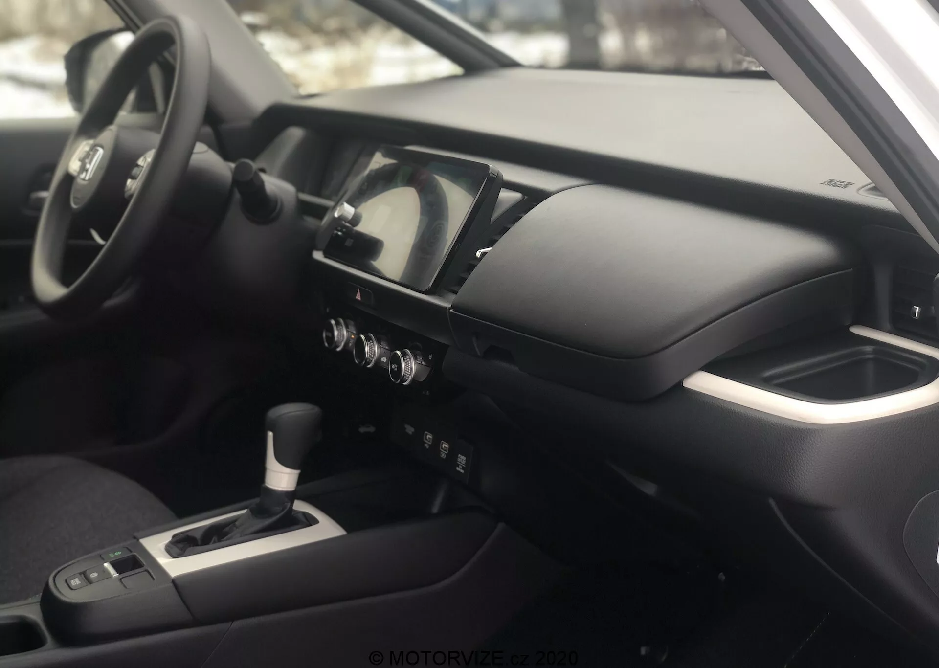 Vue intérieure de la Honda Jazz 2020 depuis le côté passager, mettant en avant le tableau de bord et le cockpit du conducteur. La photographie, prise selon un angle légèrement en hauteur, met en évidence la console centrale, le volant multifonction avec commandes intégrées, le combiné d'instruments digital-analogique, la console centrale avec levier de vitesses, les boutons de commande climatique et un écran d'info-divertissement/navigation. L'accoudoir suggère un confort de conduite, et l'intérieur dispose de matériaux métalliques et doux au toucher, reflétant une philosophie de design moderne et haut de gamme.