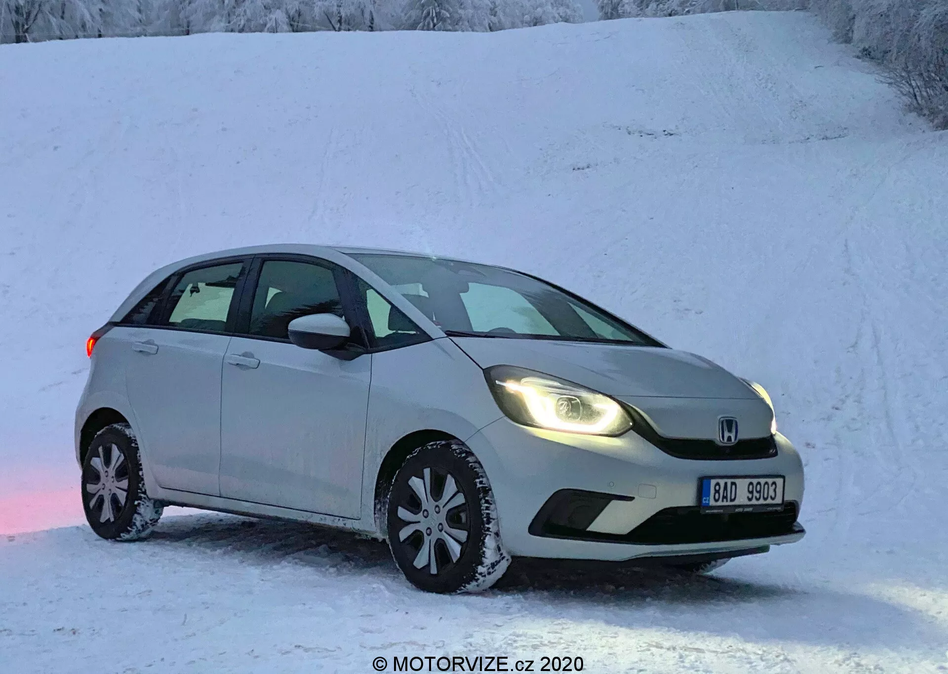 L'image présente une Honda Jazz 2020, une berline argentée ou grise clair stationnée sur une surface enneigée, évoquant une scène hivernale avec un paysage couvert de neige. Capturée sous un angle frontal trois quarts, la vue met en valeur le côté conducteur, montrant la calandre avant, les phares, le capot, le pare-brise, la porte côté conducteur et la porte arrière côté passager. La voiture est équipée de phares modernes, d'un pare-chocs avant sculpté, d'un badge proéminent sur la calandre, de rétroviseurs latéraux avec clignotants intégrés et de roues en alliage à rayons multiples dotées de pneus d'hiver. Le véhicule stationnaire est représenté sans flou de mouvement dans des conditions de faible luminosité, soit tôt le matin soit tard dans l'après-midi.