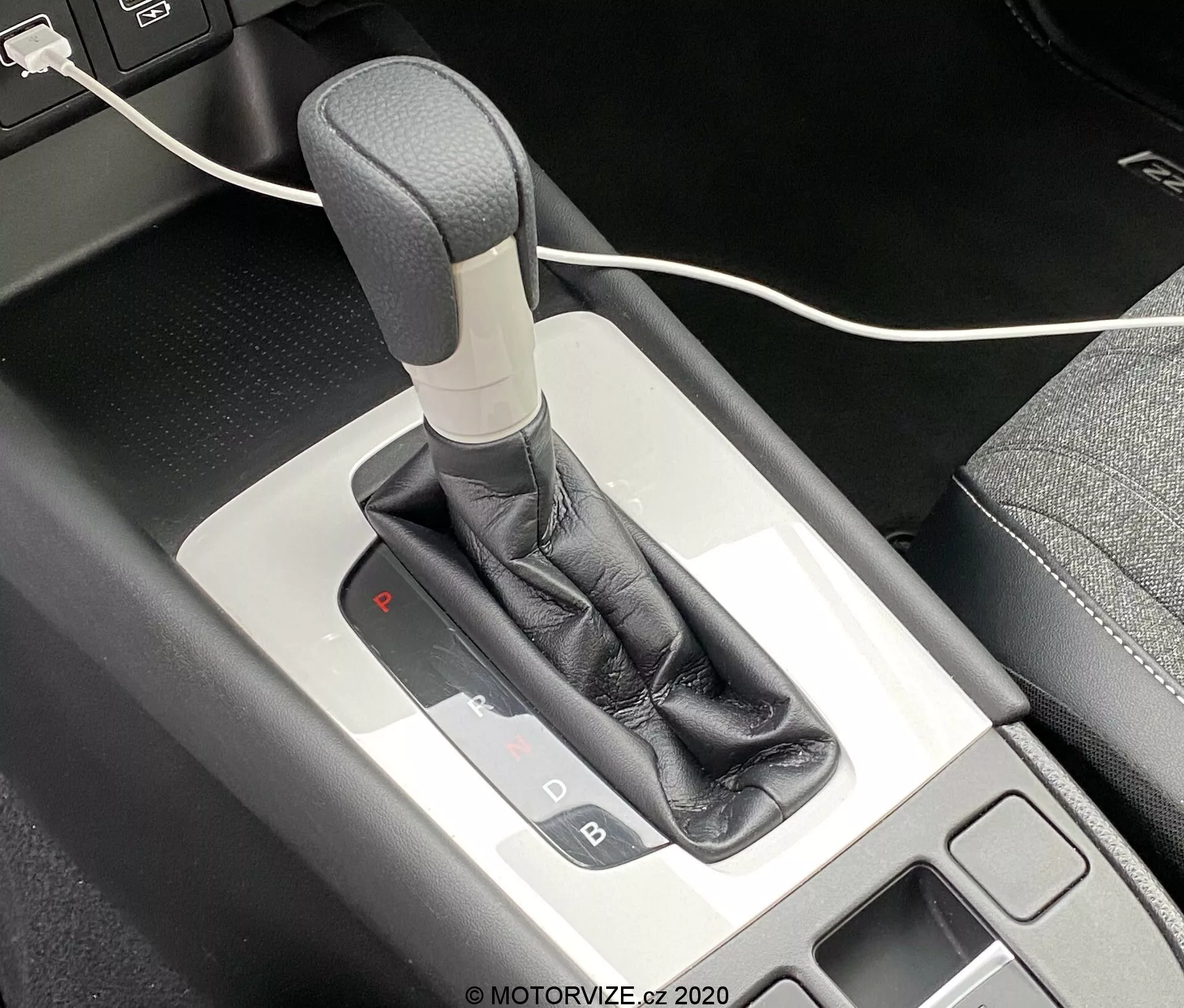 Vue rapprochée du levier de vitesses automatique de la Honda Jazz 2020 depuis un angle plongeant, montrant le mécanisme de sélection des vitesses et la console centrale environnante. Le levier de vitesses a une poignée grise ou argentée, enveloppée d'un soufflet en cuir noir, complétée par une garniture métallique. Le panneau de sélection des vitesses éclairé indique que le véhicule est en mode parking avec une lumière rouge sous le 'P'. Un petit espace de rangement avec un tapis noir est visible à gauche, ainsi qu'un câble USB partiellement visible en haut de l'image.
