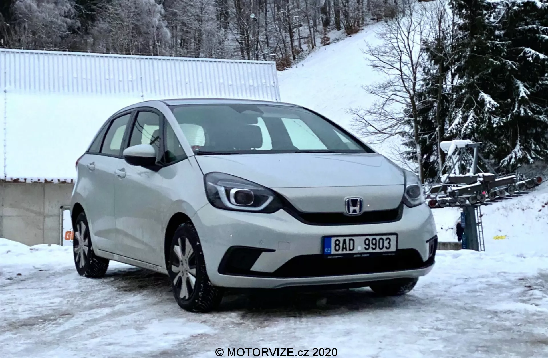 Vue de face en angle d'une Honda Jazz 2020 argentée capturée par une journée d'hiver, montrant la calandre avant, les phares, le capot et le pare-chocs avant sur un arrière-plan enneigé avec des arbres dénudés et un bâtiment blanc.