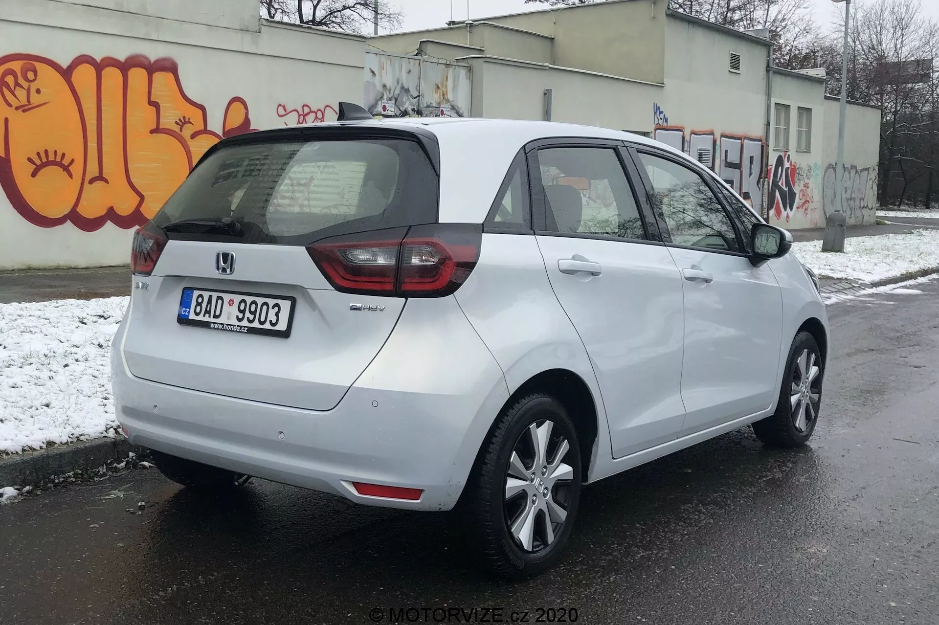 Vue arrière des trois quarts d'une Honda Jazz 2020 blanche garée sur une surface en asphalte dans un environnement urbain potentiellement froid avec de la neige au sol et des graffitis sur un mur. La voiture dispose de feux arrière mixtes LED et ampoules traditionnelles, de la plaque d'immatriculation centrale arrière et d'un badge indiquant le niveau de finition ou la variante. Elle possède un toit profilé, des jantes en alliage à cinq branches, des rétroviseurs latéraux couleur carrosserie avec indicateurs de direction intégrés et des poignées de porte assorties. Le pare-brise arrière est équipé d'un désembueur et d'un essuie-glace pour une meilleure visibilité.