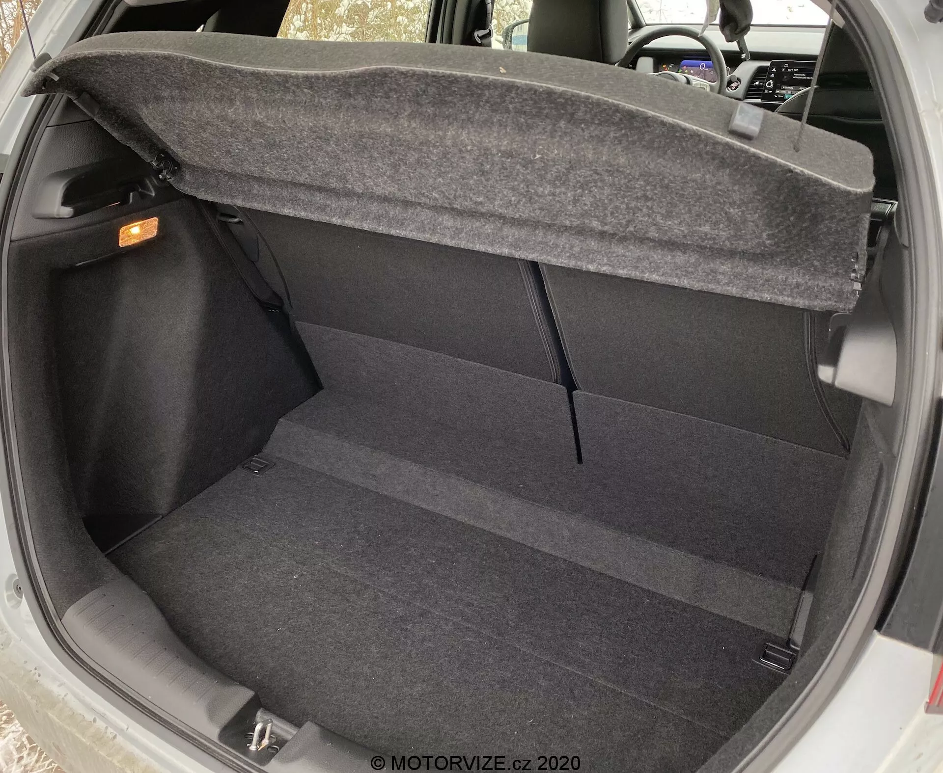 Vue intérieure de l'espace de coffre de la Honda Jazz 2020 depuis un angle légèrement surélevé, montrant le plancher propre et moquetté, les parois latérales et l'arrière des sièges arrière. Le coffre est vide, avec la poignée de déverrouillage intérieure visible dans le coin supérieur droit et le mécanisme de charnière du couvercle du coffre partiellement visible sur les côtés supérieurs, mettant en évidence la praticité et les caractéristiques de conception de base du coffre.