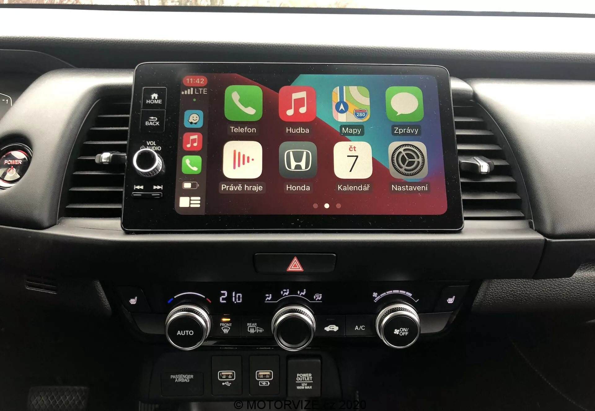 Console centrale de l'intérieur de la Honda Jazz 2020, montrant le système d'infodivertissement et le contrôle du climat depuis la perspective du conducteur. Écran tactile actif avec des applications comme Téléphone, Musique, Cartes indiquant Bluetooth, GPS, lecture multimédia et Apple CarPlay. Sous l'écran, boutons physiques, feux de détresse et cadrans de contrôle climatique à double zone avec lectures en Celsius.