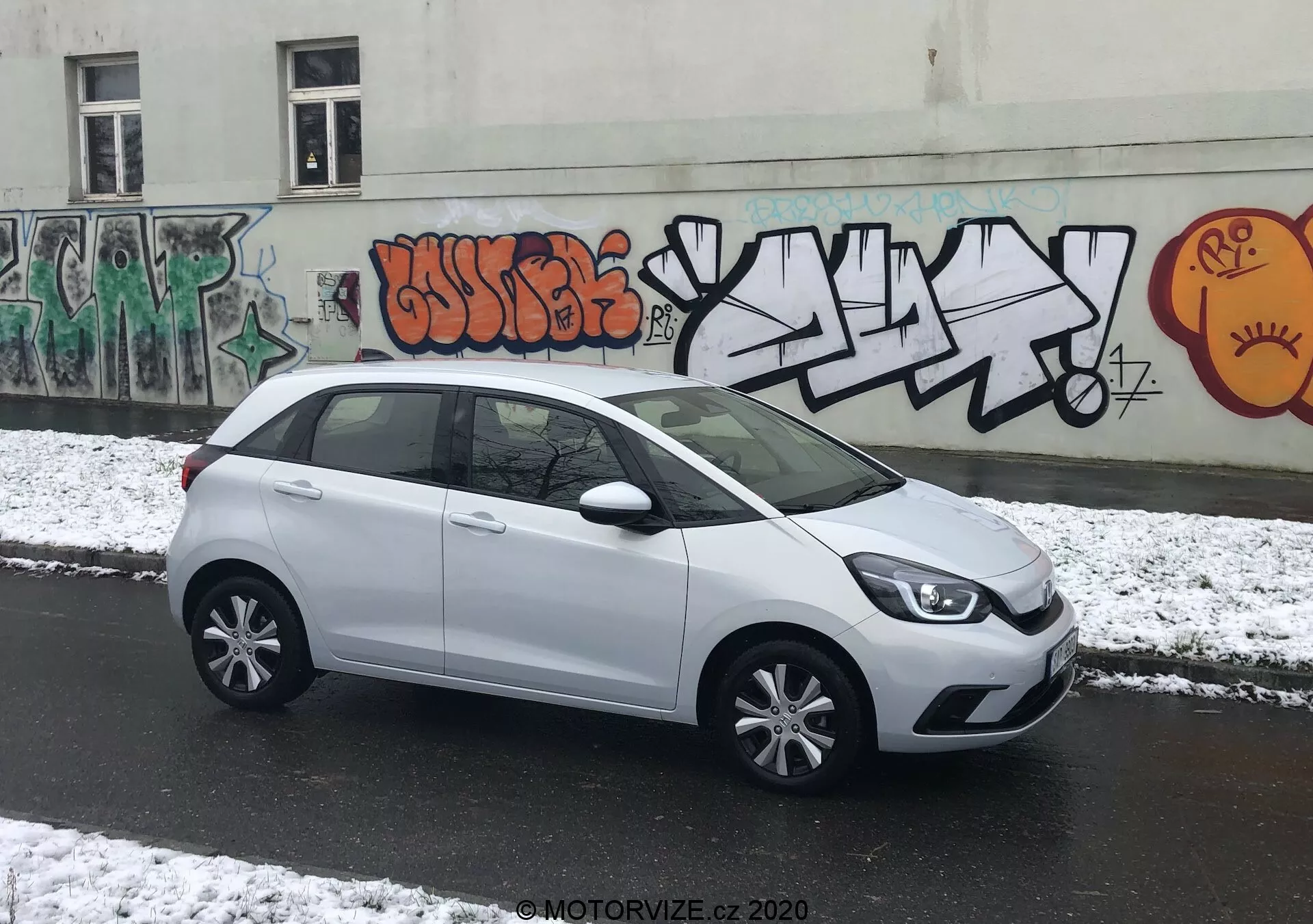 L'image présente une Honda Jazz 2020 blanche, stationnée perpendiculairement à la caméra dans une rue urbaine, permettant une vue latérale complète. L'avant élégant de la voiture avec ses blocs optiques modernes est partiellement visible, menant à une ligne de caractère douce sur le côté. Les poignées de portes assorties à la carrosserie, les rétroviseurs latéraux et les enjoliveurs à rayons multiples accentuent son apparence élégante. La vaste surface vitrée offre beaucoup d'espace pour la tête, et une petite vitre de custode arrière ajoute au design. Le spoiler arrière suggère une efficacité aérodynamique. L'éclairage nuageux avec de la neige fraîche et le fond urbain graffité ajoute un environnement dynamique.