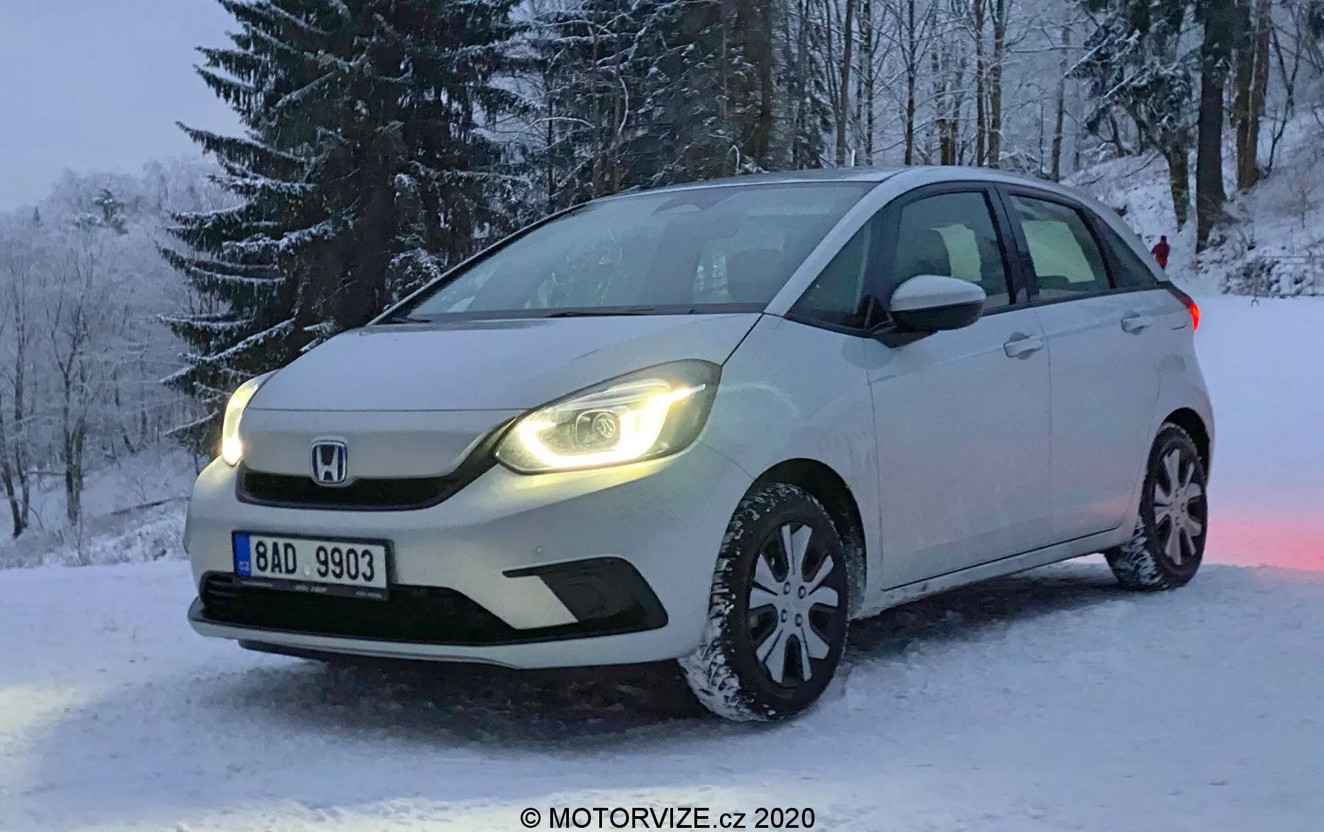 Photographie professionnelle d'une Honda Jazz blanche de 2020, stationnée sur un terrain enneigé en hiver. L'image est prise sous un angle qui met en avant le côté avant gauche de la voiture, montrant la calandre, les phares à LED, le capot, la roue avant gauche à multiples rayons équipée de pneus neige, le pare-brise et la porte conducteur. Des arbres couverts de neige sont visibles en arrière-plan et la plaque d'immatriculation est lisible. La faible luminosité suggère le matin ou la fin d'après-midi, avec les feux de jour de la voiture se reflétant sur la neige, renforçant l'ambiance de la scène. Le véhicule dispose d'un éclairage avancé et de clignotants intégrés dans les rétroviseurs latéraux, avec le badge distinctif Honda affiché de manière bien visible sur la calandre.