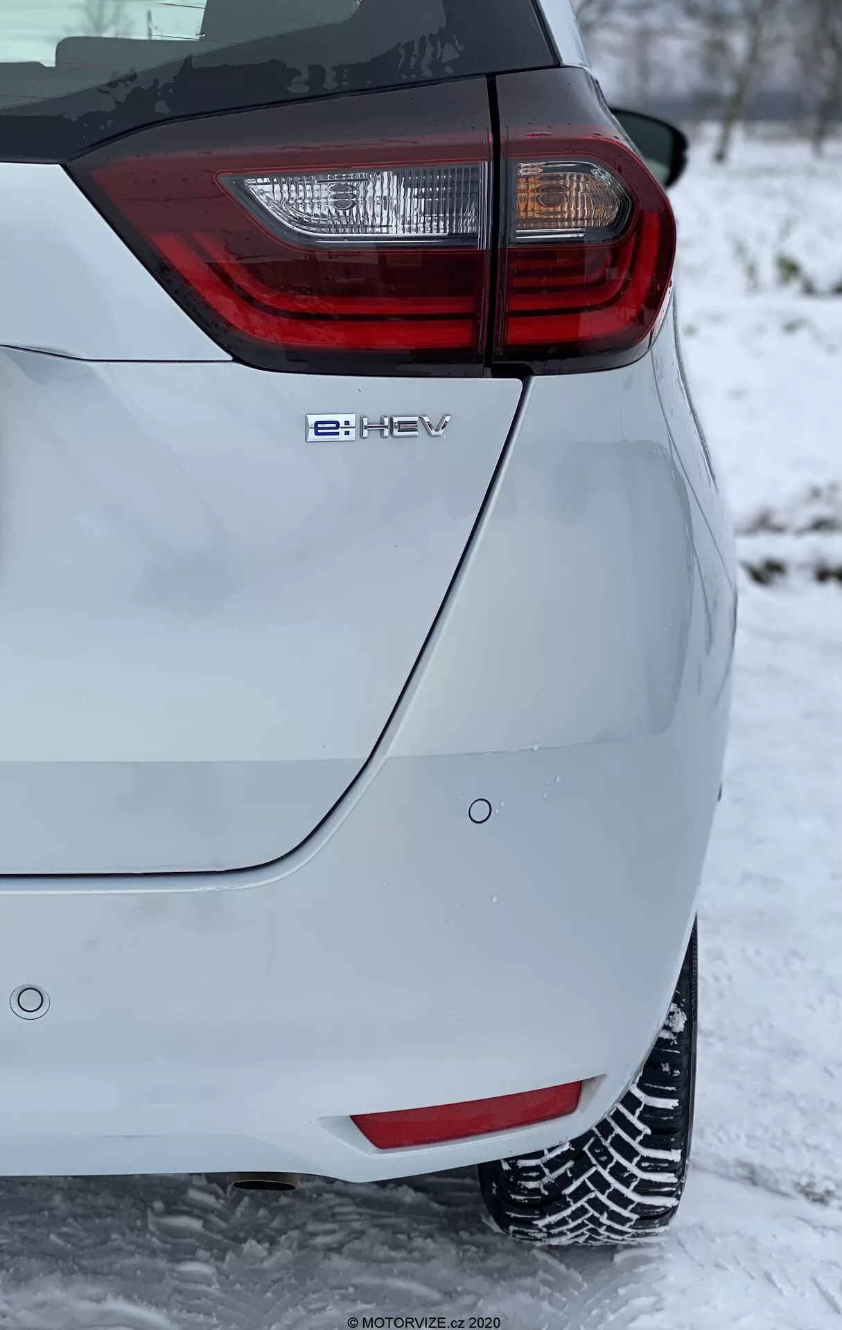 Gros plan sur le coin arrière d'une voiture à hayon moderne, présentant un groupe de feux arrière proéminent avec des éléments rouges et transparents. L'image, prise en orientation verticale, montre l'emblème de la technologie hybride 'e-HEV' et se déroule dans un environnement hivernal avec de la neige au sol et recouvrant les pneus, indiquant des conditions de conduite hivernales. La peinture de couleur claire du véhicule contraste avec le décor enneigé, mettant en évidence ses caractéristiques de design moderne et écologique.