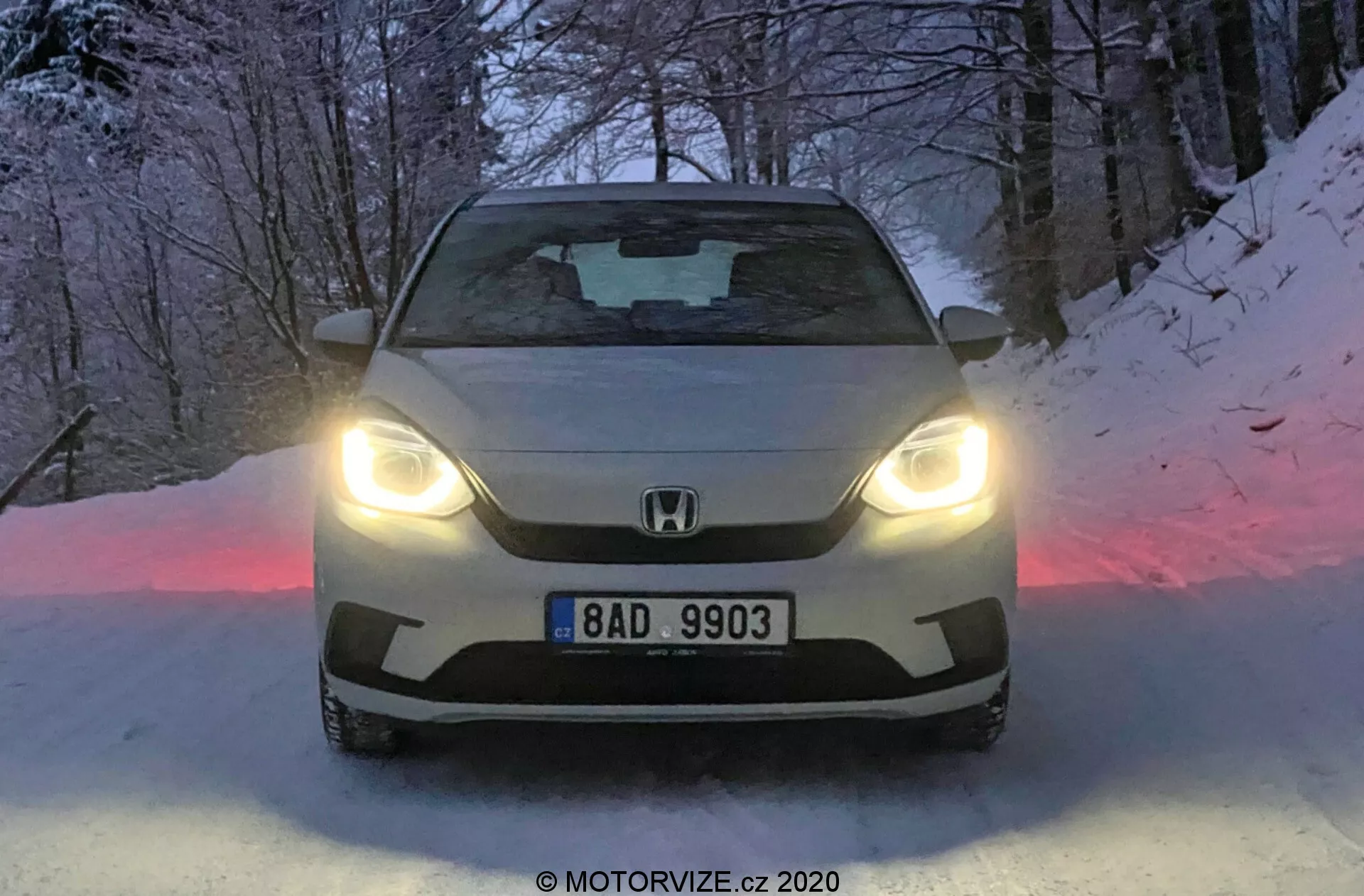Prise de vue frontale de la Honda Jazz 2020 avec phares LED, affichant le logo de la marque et la plaque d'immatriculation sur la calandre, avec un capot sculpté et un pare-brise teinté. Le sol enneigé et l'ambiance crépusculaire rehaussent le design avancé et les capacités de conduite hivernale de la voiture.