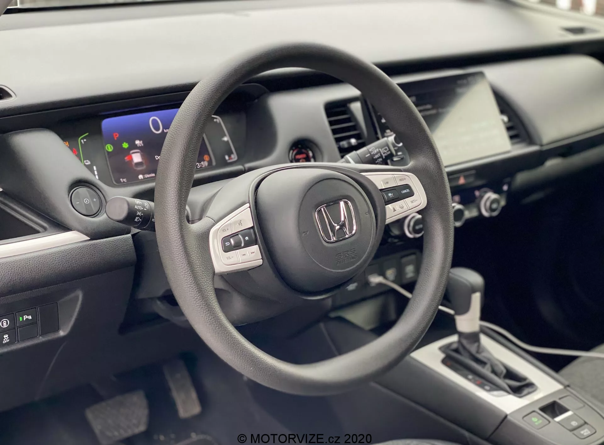 La photo montre l'intérieur d'une Honda Jazz 2020 vu de la perspective du conducteur. L'accent est mis sur le volant avec le logo Honda, équipé de plusieurs boutons de contrôle suggérant des fonctionnalités audio intégrées, un affichage d'informations et potentiellement un régulateur de vitesse. Derrière le volant, un affichage numérique partiel montre un indicateur de niveau de batterie, ce qui laisse penser que le véhicule pourrait être hybride ou entièrement électrique. L'intérieur bien éclairé et moderne comprend également une console centrale avec un système d'infodivertissement tactile, des commandes de climatisation et un levier de vitesses à design contemporain, indiquant une transmission automatique. L'image met en évidence les finitions mate, satinée et brillante utilisées dans les matériaux de l'intérieur de la voiture, soulignant le design ergonomique et riche en technologie du véhicule.