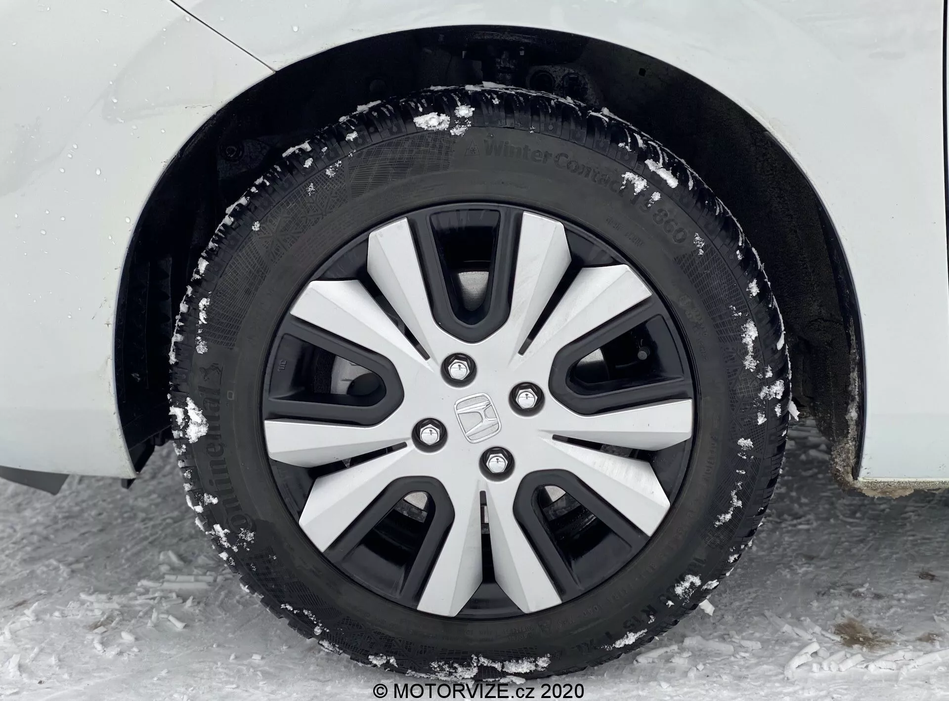Vue rapprochée de la roue avant gauche d'une Honda Jazz 2020, capturée sous un angle bas légèrement en avant de la roue et orientée vers l'arrière. La jante en alliage à cinq branches présente un design argenté et noir, recouverte d'une fine couche de neige et de petites particules de glace, suggérant des conditions hivernales. Le pneu semble être une variété hivernale avec des motifs de bande de roulement spécialisés pour les conditions de neige et de glace. Le garde-boue de la voiture est partiellement visible et la carrosserie semble être blanche avec un revêtement en plastique noir dans le passage de roue.