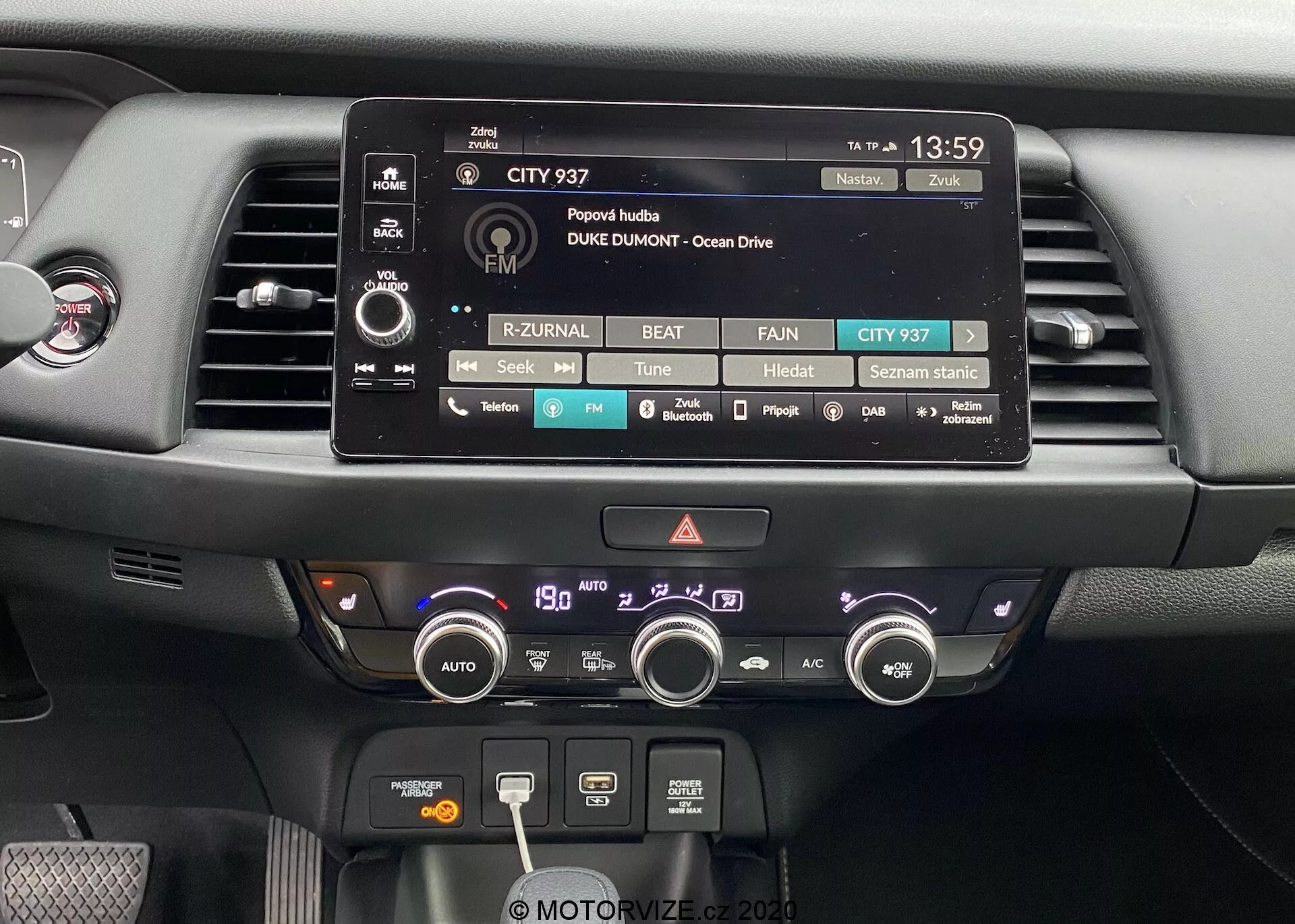 Vue rapprochée de la zone du tableau de bord central de la Honda Jazz 2020. L'image présente en évidence l'écran tactile du système d'infodivertissement, entouré de boutons et de molettes physiques pour les réglages audio, médias et du véhicule. En dessous se trouvent deux aérations et un système de contrôle climatique comprenant des boutons et des molettes pour ajuster la température, la vitesse du ventilateur, les fonctions de dégivrage et la recirculation. Le tableau de bord est composé de matériaux modernes, incluant des plastiques mats et brillants, conçu avec une accessibilité fonctionnelle et une facilité d'utilisation à l'esprit.