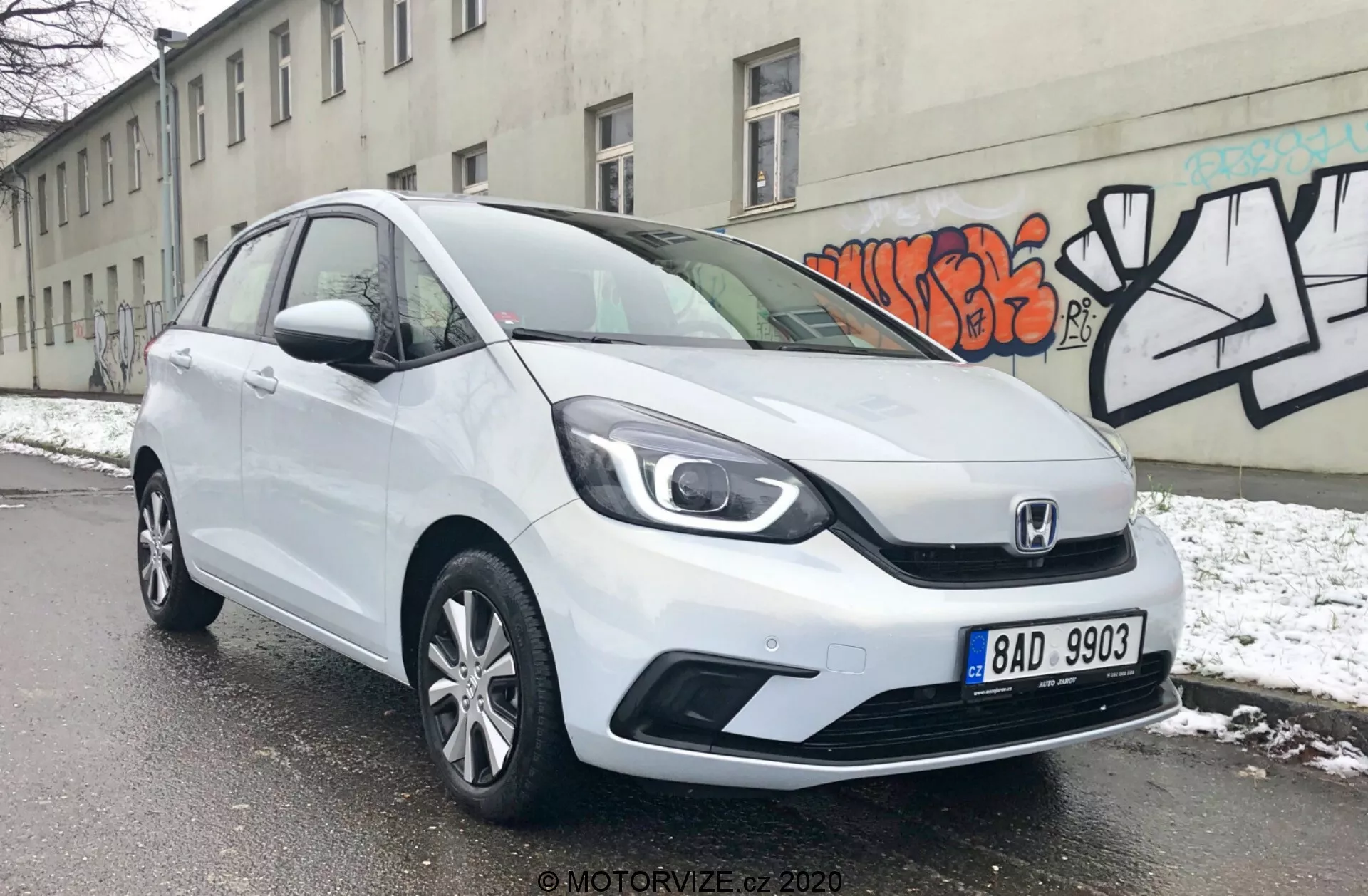 L'image montre un véhicule hatchback Honda Jazz 2020 blanc sous un angle trois-quarts avant, révélant ses phares élégants, la calandre distinctive de Honda avec un design à deux barres et les lignes de contour aérodynamiques sur le capot. Le pare-chocs avant présente des prises d'air visibles et possiblement des phares antibrouillard, tandis que le profil latéral montre des roues en alliage à plusieurs branches et des panneaux de porte légèrement contourés. La voiture est stationnée dans une rue enneigée, indiquant un environnement hivernal, avec un mur gris décoré de graffitis en arrière-plan, soulignant un cadre urbain.