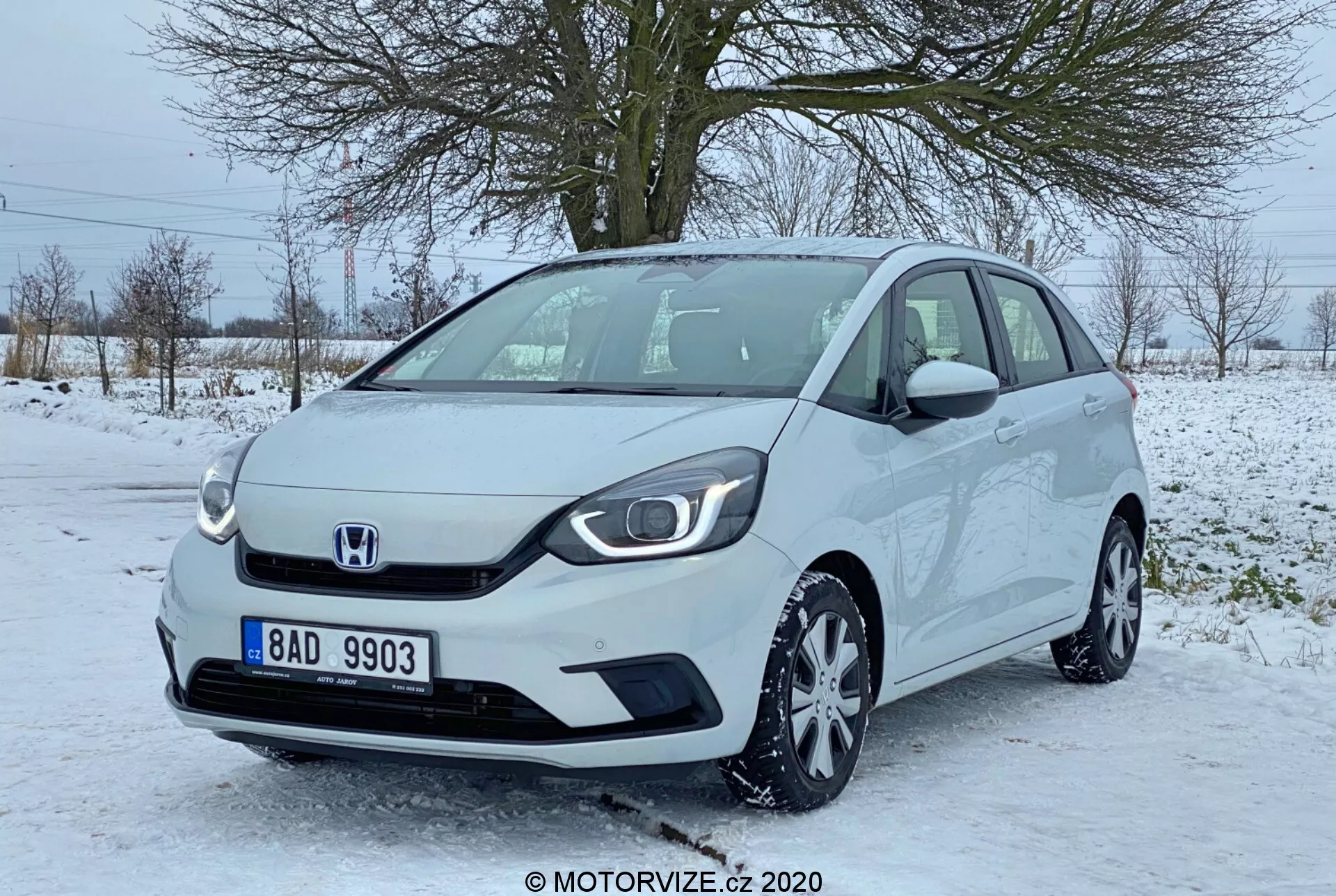 Vue de trois quarts avant d'une Honda Jazz 2020, montrant la partie avant et une partie du côté passager de cette compacte moderne. L'image, apparemment prise à hauteur d'œil, met en évidence la calandre minimaliste avec un emblème central Honda, des phares élégants avec des feux de jour à LED, des jantes en alliage multi-rayons et des rétroviseurs latéraux avec clignotants intégrés. La voiture est garée sur une zone pavée partiellement couverte de neige, suggérant un cadre hivernal avec des arbres sans feuilles et un ciel gris en arrière-plan. Le véhicule semble propre et bien entretenu.