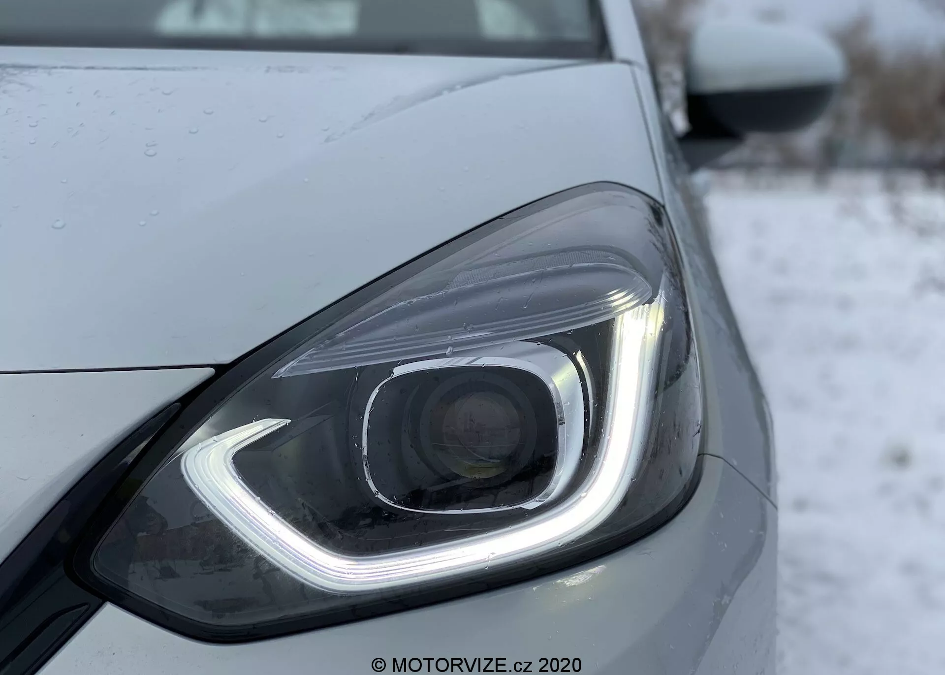 Photographie en gros plan du phare avant gauche d'une Honda Jazz 2020, mettant en valeur son design moderne avec des éléments d'éclairage LED ou HID. Le phare se distingue par une lumière de jour en forme de crochet, une lentille de projecteur et des gouttelettes d'eau suggérant des précipitations récentes ou un lavage. Le design élégant est accentué par des lignes et des contours lisses, avec un fond froid ou hivernal, possiblement enneigé.