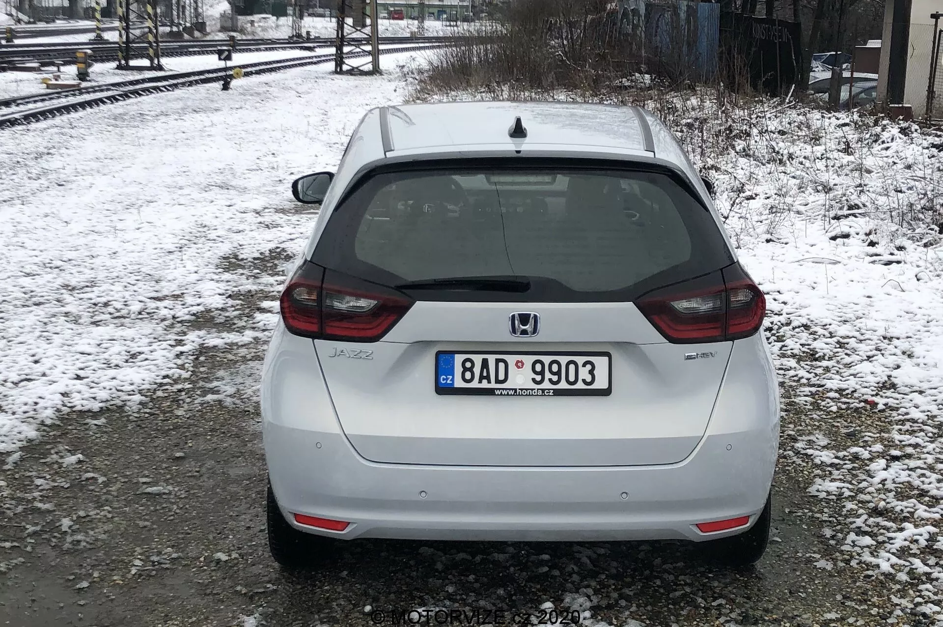 L'image montre la vue arrière d'une Honda Jazz 2020 blanche, garée sur un sol enneigé près des voies ferrées sous des conditions de lumière diffuse ou nuageuse. La voiture occupe la majorité du cadre, avec une vue arrière centrée montrant sa plaque d'immatriculation de style européen, l'essuie-glace arrière, le feu stop haut monté et l'emblème Honda. Le badge du modèle 'JAZZ' est visible sur la partie inférieure gauche du hayon, et il y a une caméra de recul au-dessus de la plaque d'immatriculation. Les feux arrière enveloppent partiellement les côtés, avec des réflecteurs sur le bas du pare-chocs, qui a également une section noire intégrée. Les signes de saleté suggèrent que la voiture a été conduite sur des routes mouillées ou boueuses. La neige et les arbres nus indiquent qu'il est hiver ou début du printemps.