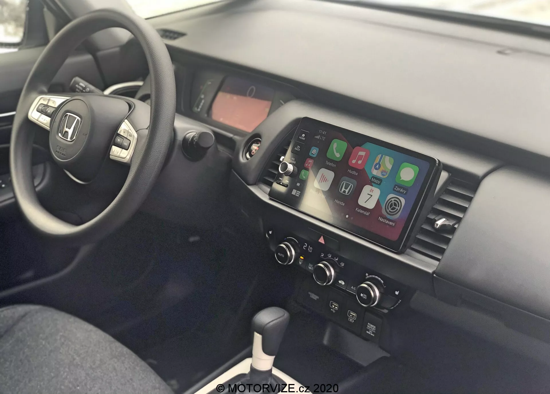 Vue intérieure d'une Honda Jazz 2020, montrant la zone du conducteur et la console centrale depuis le côté passager. Le volant est à gauche et comprend des commandes intégrées pour l'audio, la connectivité téléphonique et le régulateur de vitesse, indiquant un design pour la conduite à droite. La console centrale est équipée d'un écran tactile affichant l'intégration de smartphones comme Apple CarPlay ou Android Auto, de commandes manuelles de la climatisation, d'un levier de vitesses pour transmission manuelle, de ports USB et d'une prise électrique 12V, mettant en évidence la connectivité moderne et le design fonctionnel.