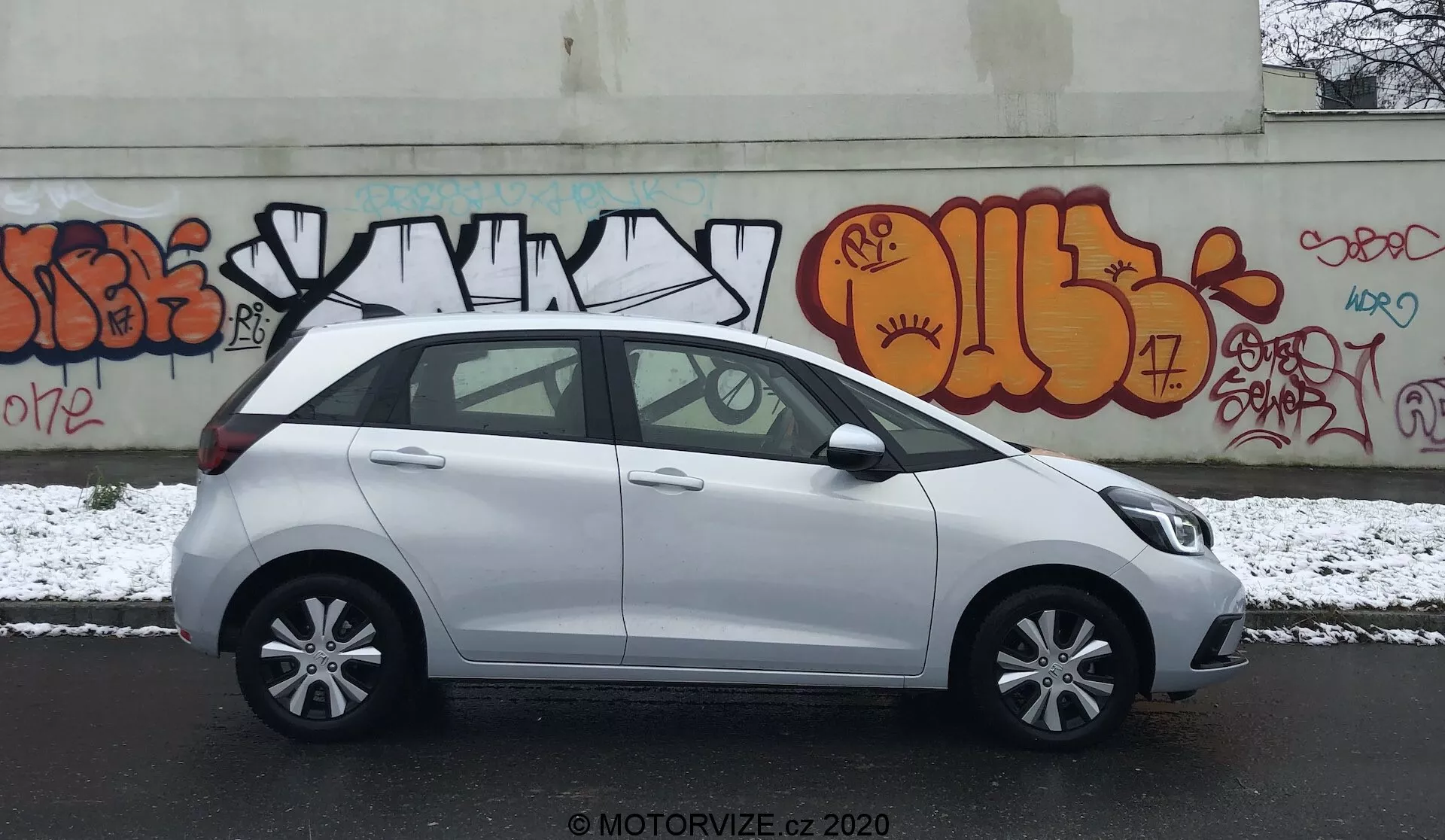 Profil latéral d'une Honda Jazz compacte blanche de 2020, garée dans une rue urbaine avec des graffitis sur le mur en arrière-plan. La scène se déroule par un jour couvert avec une légère couche de neige au sol, suggérant des conditions hivernales. L'image capture le côté conducteur, montrant des poignées de porte de la même couleur que la carrosserie, des rétroviseurs latéraux, des jantes en alliage à cinq branches et des fenêtres standard avec un montant B noir. Le véhicule semble propre avec quelques taches humides dues à la neige. Le filigrane 'MOTORVIZE.CZ 2020' est visible, indiquant la source de la photo.