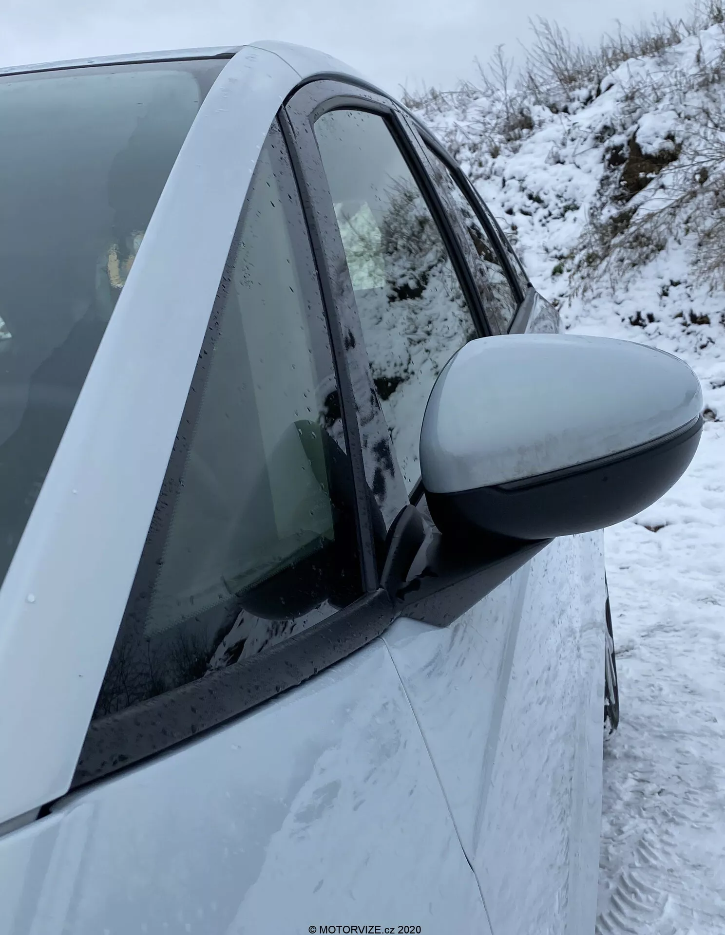 Gros plan sur le côté droit d'une Honda Jazz 2020 montrant la zone de la porte du conducteur. Le rétroviseur latéral proéminent s'étend de la gauche de l'image jusqu'au centre, indiquant une utilisation récente ou proche. Le boîtier du rétroviseur correspond à la couleur de la voiture, suggérant qu'il s'agit d'une caractéristique standard. Les éléments de design moderne incluent un rétroviseur aérodynamique et une carrosserie intacte. La fenêtre est levée avec de la condensation à l'intérieur et des flocons de neige sont visibles sur la porte et la fenêtre. Le montant B visible et des parties des deux portes, avant et arrière, indiquent un véhicule à quatre portes. L'indicateur latéral n'est pas visible. L'arrière-plan montre des conditions hivernales avec de la neige au sol.