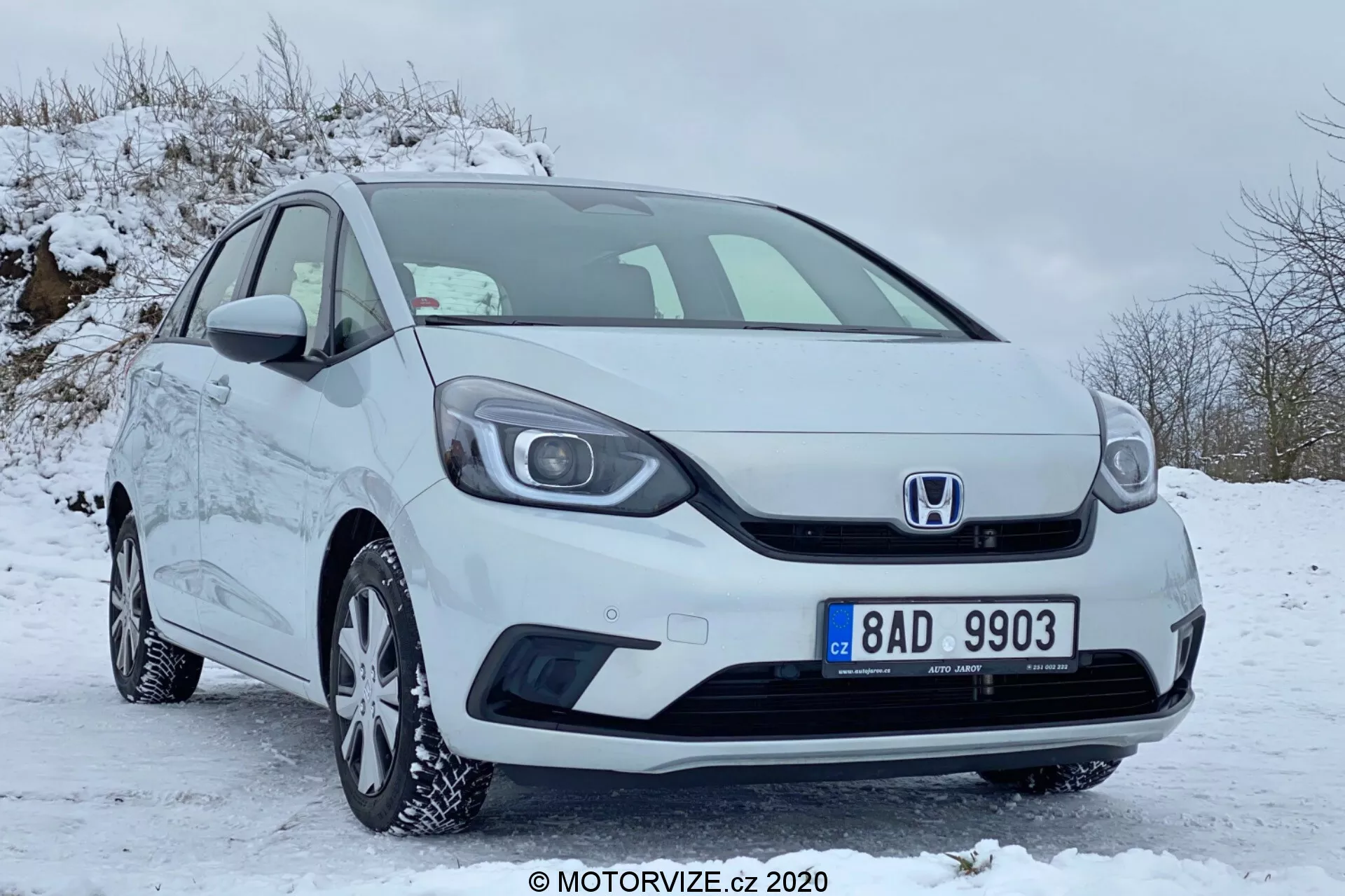 Vue de trois quarts avant d'une Honda Jazz compacte blanche de 2020, garée dans une zone couverte de neige, mettant en valeur la calandre avec l'emblème Honda, le pare-chocs avant, les phares, les jantes en alliage avec pneus hiver, les rétroviseurs latéraux et les poignées de porte, le tout en bon état et bien entretenu.