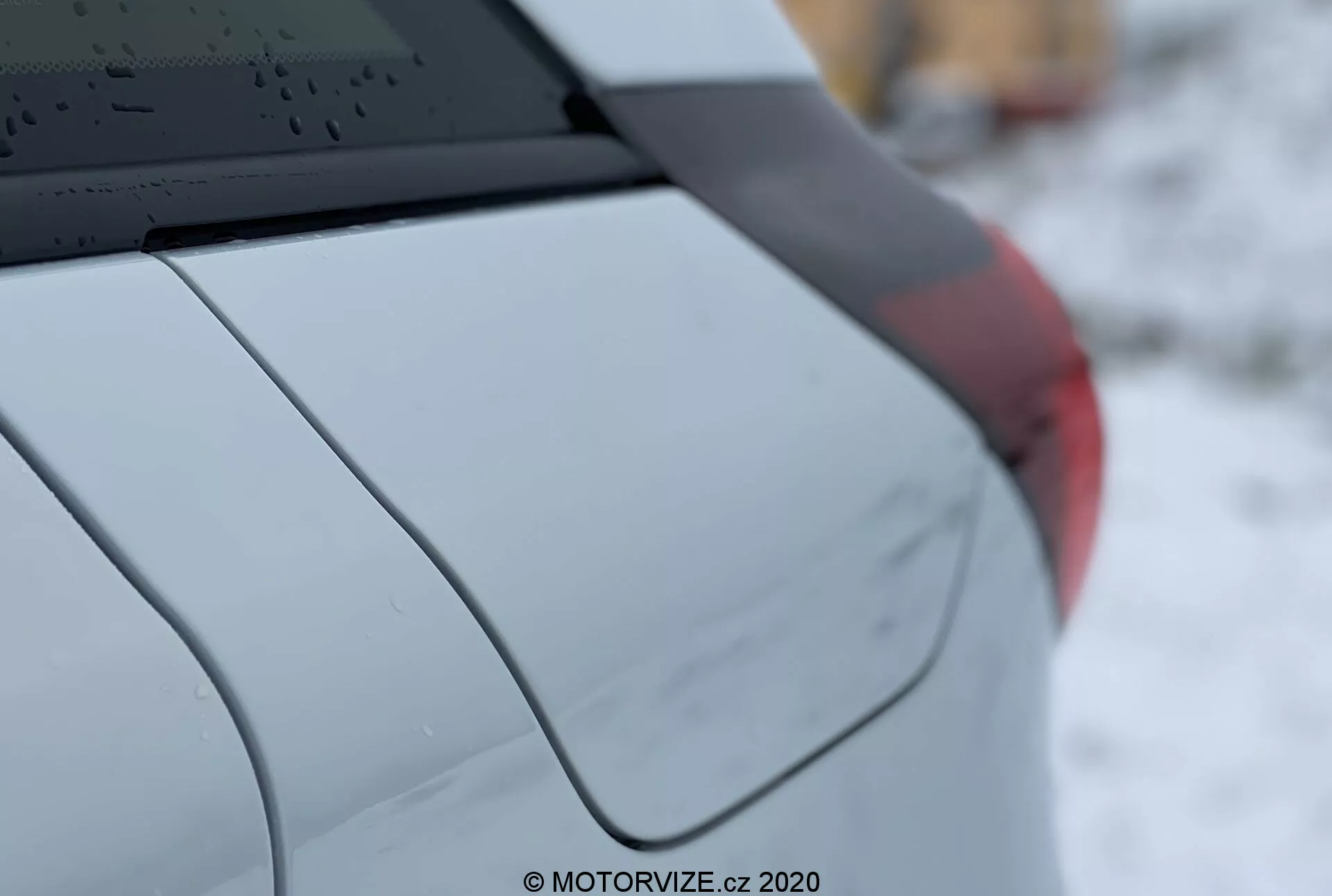 Gros plan sur la Honda Jazz 2020, montrant le quart arrière droit de la voiture vue de côté à trois quarts. Les caractéristiques comprennent une vitre arrière avec des lignes de dégivrage, une antenne en aileron de requin montée sur le toit, le montant C accentuant le design de la voiture, un garde-boue arrière avec une courbure lisse, un fragment de l'assemblage des phares arrière et une finition de peinture métallisée ou nacrée. La faible profondeur de champ met en évidence les détails du véhicule tandis que l'arrière-plan est flou. Le filigrane indique 'MOTORVIZE.cz, 2020.'