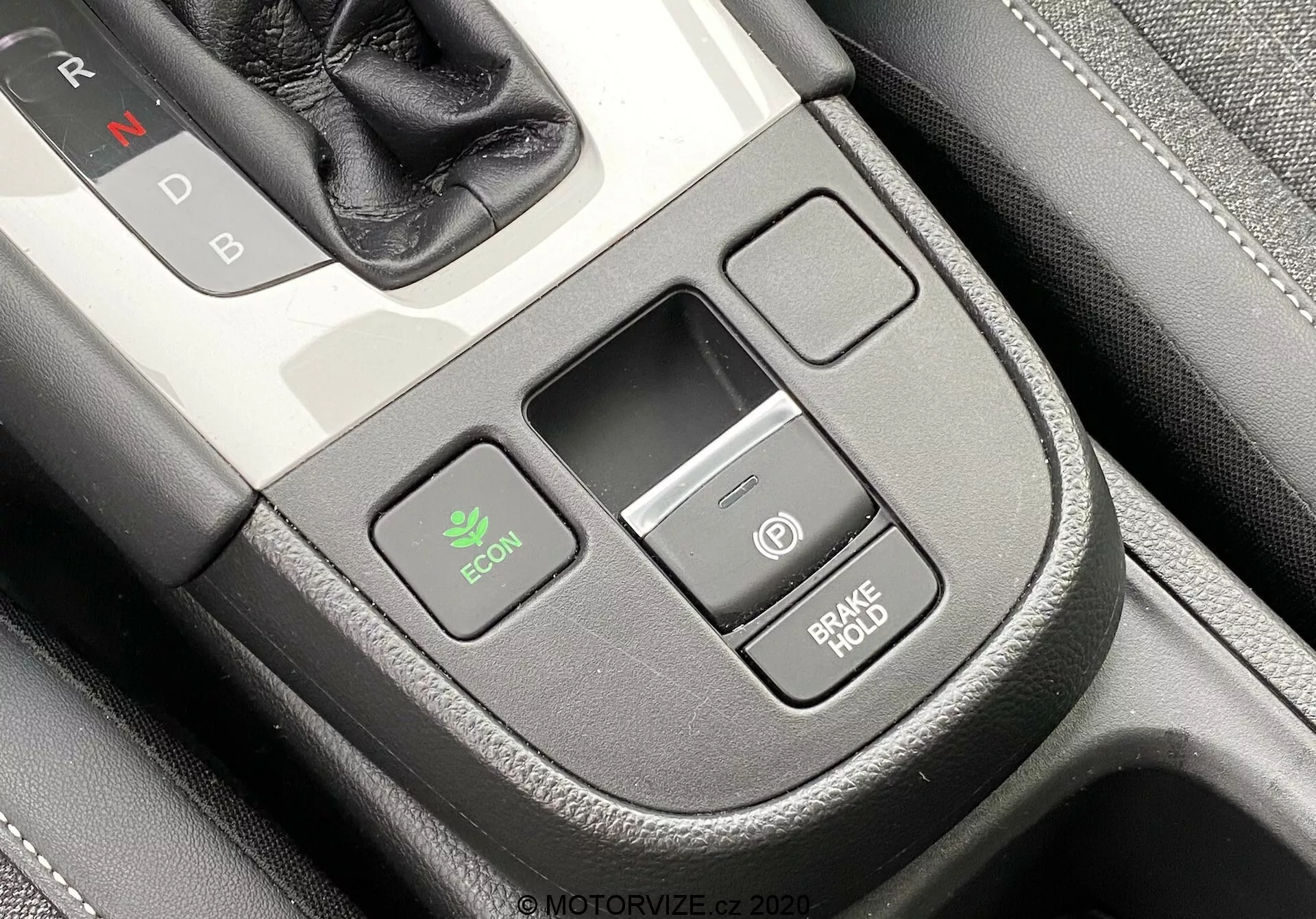 Gros plan de la console centrale intérieure d'une Honda Jazz 2020 vue légèrement en plongée depuis la perspective du conducteur. Le sélecteur de vitesse avec 'N' pour Neutre est partiellement visible en haut à droite, indiquant une transmission automatique. On remarque le bouton ECON à gauche pour le mode économique, une petite fente de rangement rectangulaire au centre et deux boutons empilés à droite—un avec une icône d'alimentation et l'autre marqué 'BRAKE HOLD,' qui évite que la voiture roule à l'arrêt. La console a une finition de qualité avec une texture similaire au cuir et un détail de couture.