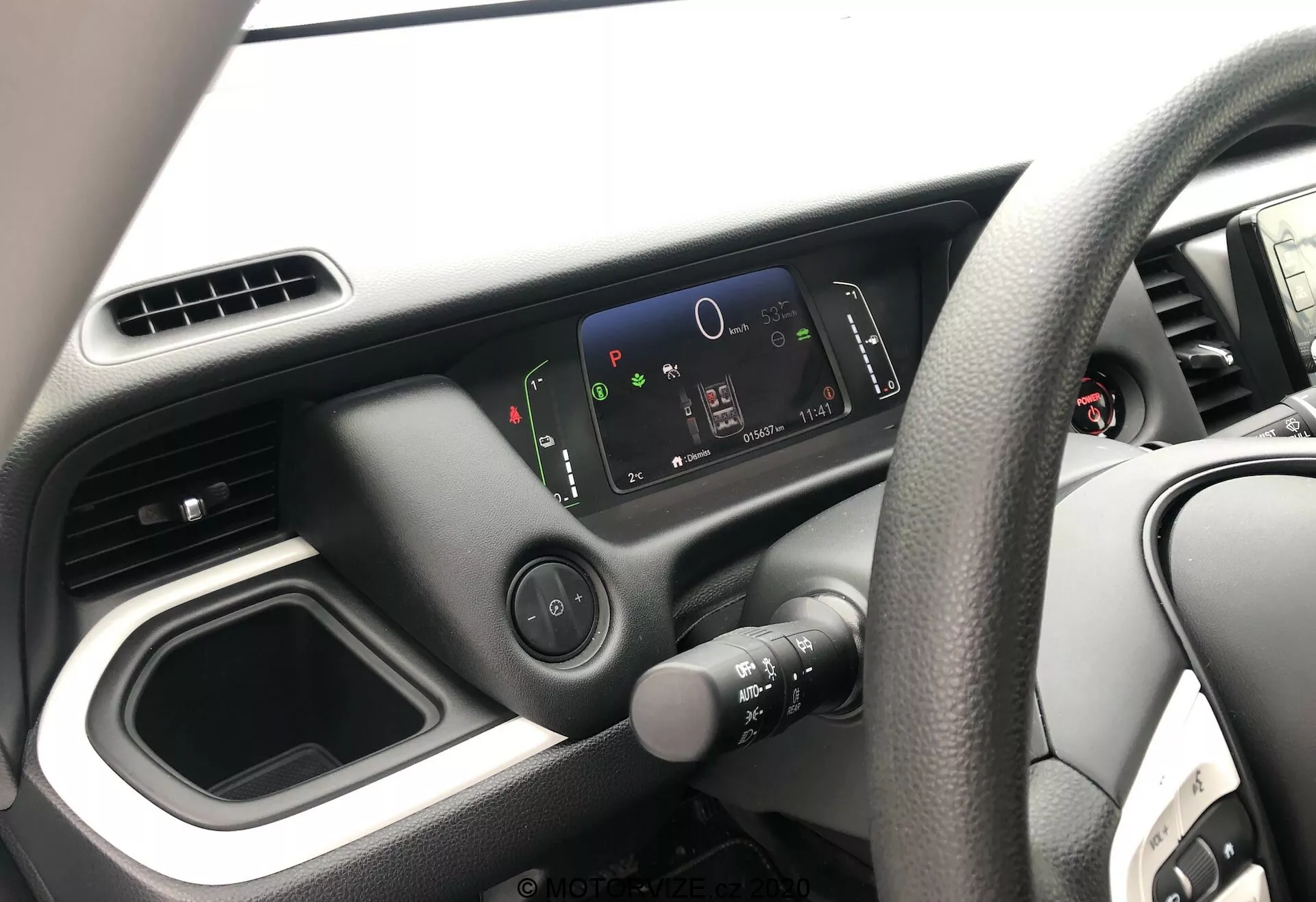 Vue intérieure de la Honda Jazz 2020, se concentrant sur le côté du conducteur depuis la perspective du passager. L'image présente le volant à droite avec un design à quatre branches, le tableau de bord minimaliste avec un écran numérique haute résolution et la disposition simple de la console centrale avec un compartiment de rangement vide et des bouches d'aération. Seule une partie de la porte du conducteur est visible.