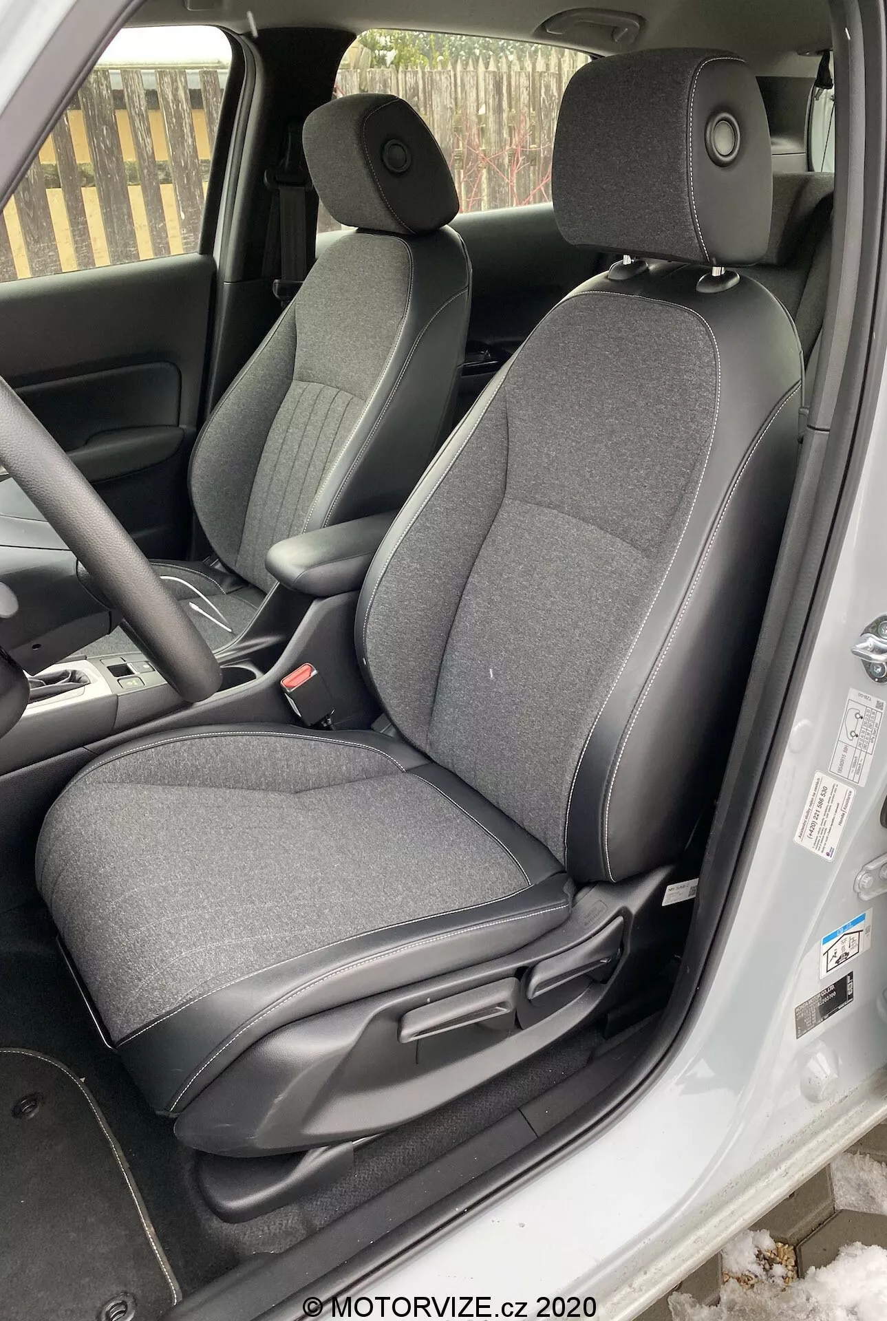 L'image montre l'intérieur d'une Honda Jazz 2020, se concentrant sur la zone du siège conducteur, prise d'un angle légèrement au-dessus de la ligne des yeux depuis l'extérieur du véhicule. Le siège conducteur ajustable est recouvert d'un tissu sombre. Sont visibles les leviers de réglage pour glisser et incliner le siège, le panneau intérieur de la porte avec les commandes des vitres électriques, le volant partiellement visible avec des contrôles montés sur le volant, une partie du tableau de bord, le tapis de sol et la zone des pédales indiquant une possible transmission manuelle, une partie de la console centrale, et la ceinture de sécurité du conducteur pendante sur le côté.