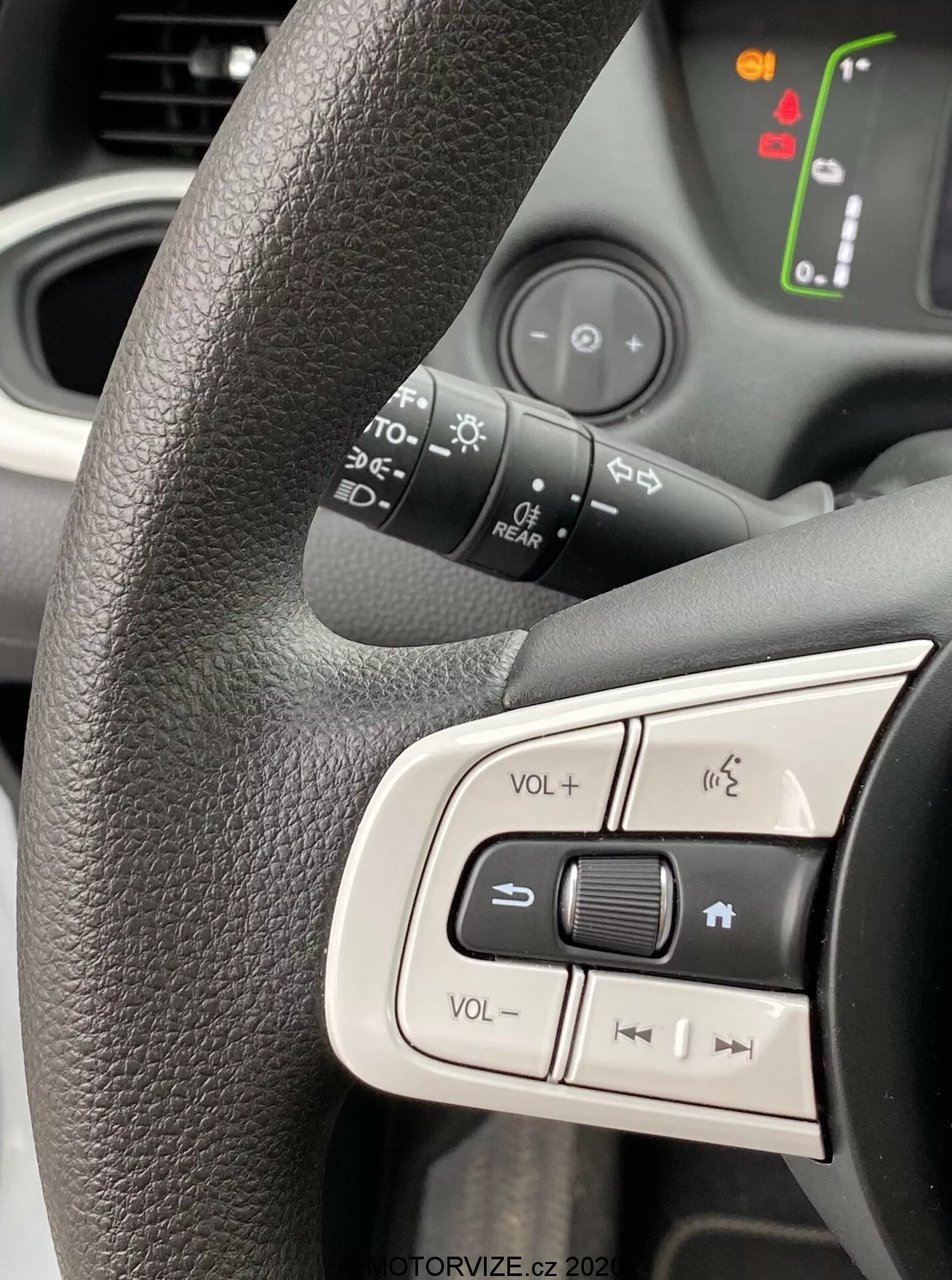 Vue rapprochée du côté gauche du volant d'une Honda Jazz 2020, mettant en avant les boutons de commande intégrés. Volant noir texturé avec une prise ergonomique, affichant les contrôles de volume, le bouton de sourdine et les boutons de navigation multimédia. L'arrière-plan légèrement flou révèle des symboles illuminés sur le tableau de bord, indiquant que le véhicule est allumé. La lumière naturelle capturant le design moderne et convivial, accentuant l'ergonomie intérieure et les fonctionnalités technologiques.