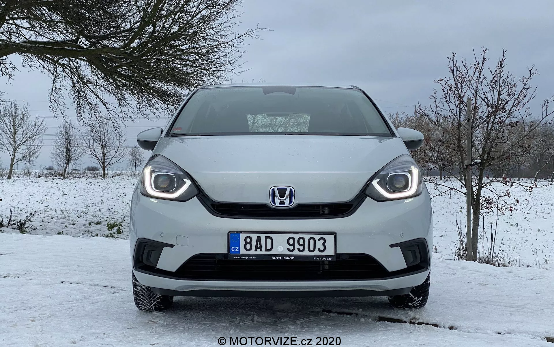 Vue de face d'une Honda Jazz 2020 en conditions hivernales, mettant en avant un design de calandre distinctif, des phares visibles avec feux de jour à LED et une plaque d'immatriculation centrale, stationnée sur une route enneigée avec un ciel couvert et des arbres sans feuilles.