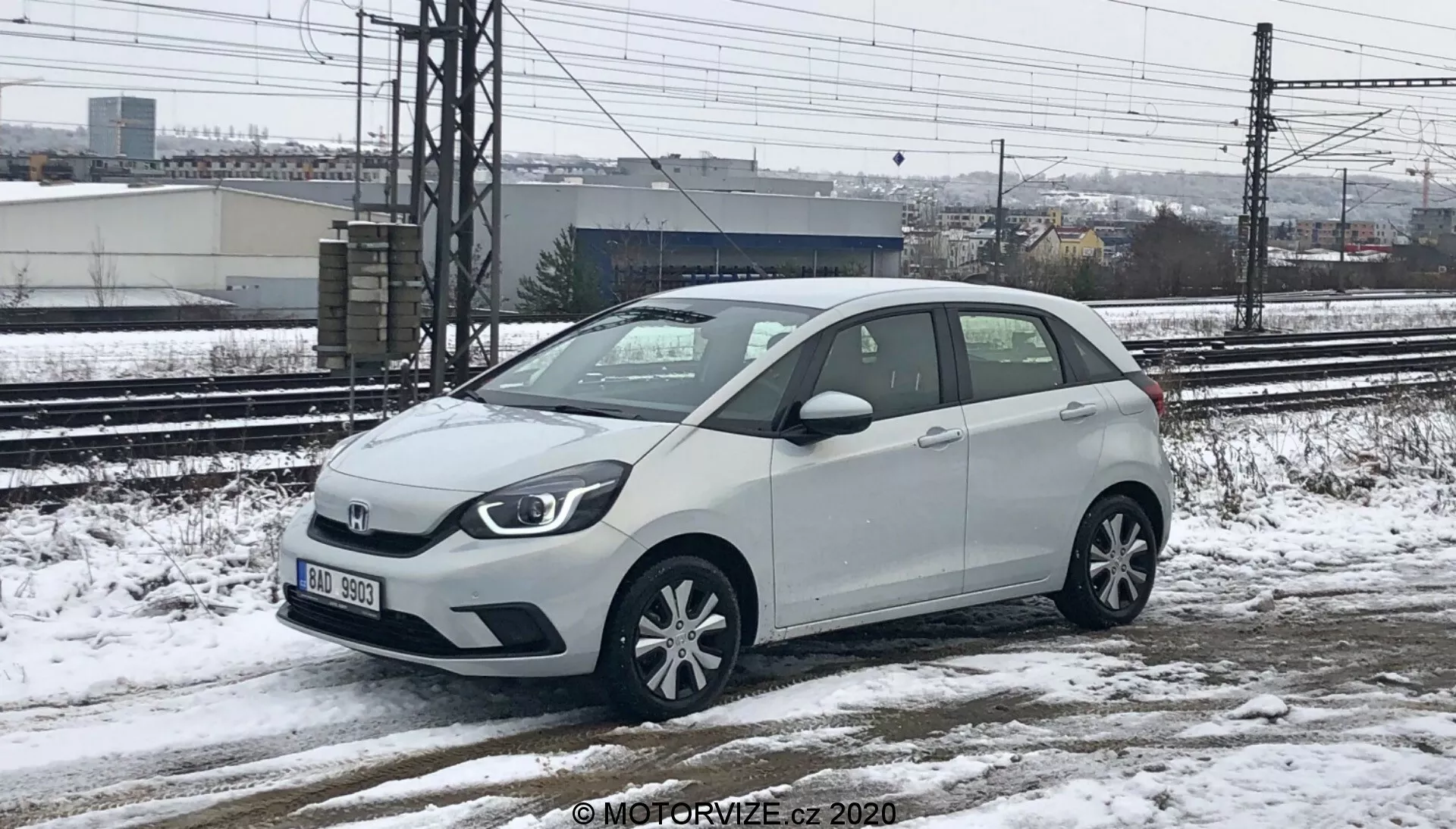 Une Honda Jazz 2020 blanche, une voiture compacte de catégorie hatchback, capturée d'une perspective de hauteur basse à moyenne, montrant à la fois l'avant et le côté droit. La voiture est garée sur une surface enneigée et mouillée, avec de la neige visible sur le véhicule et le sol, indiquant des conditions météorologiques froides. Des voies ferrées et des structures industrielles sont visibles en arrière-plan sous un ciel couvert. La voiture présente une calandre avant principale avec l'emblème de la marque, des phares halogènes, des jantes en alliage à plusieurs rayons, des rétroviseurs latéraux avec clignotants intégrés et une courte antenne sur le toit arrière.