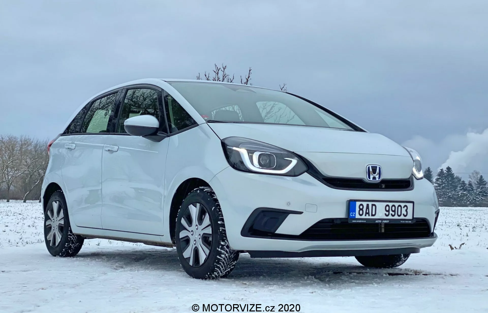 L'image présente une Honda Jazz 2020 compacte blanche, placée dans un cadre hivernal avec un sol enneigé et un ciel partiellement nuageux. La voiture est vue sous un angle de quart avant axé sur le côté conducteur, mettant en évidence la grille distinctive avec le logo au centre, les phares LED élégants et le pare-chocs avant lisse avec un logement intégré pour les phares antibrouillard. Le profil latéral montre une ligne de toit inclinée, des roues en alliage multi-branches et des rétroviseurs et poignées de portes assortis à la couleur de la carrosserie. L'état impeccable du véhicule suggère qu'il pourrait s'agir d'une image promotionnelle ou d'une revue capturée en 2020 par MOTORVIZE.cz.