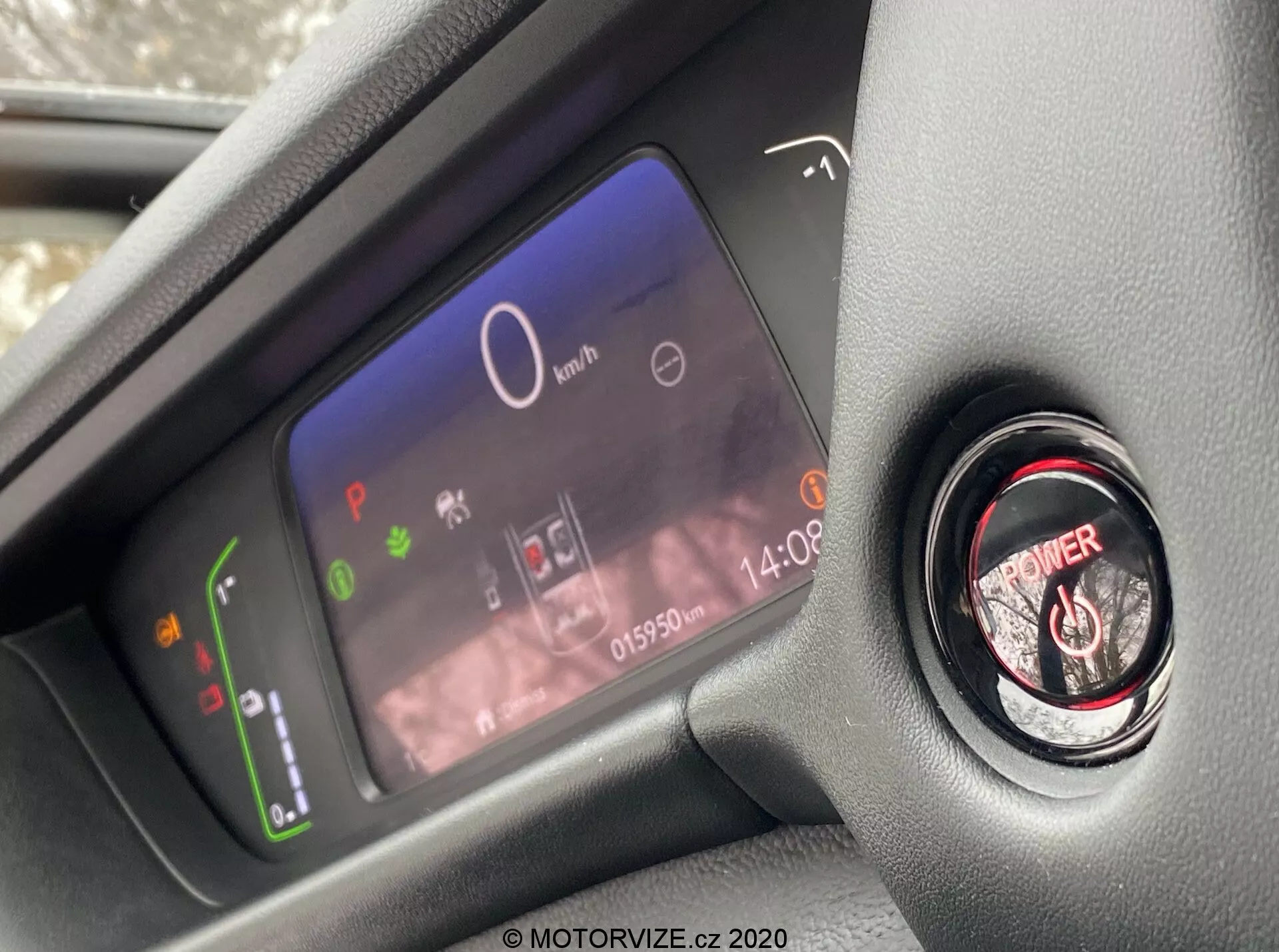 Vue rapprochée du tableau de bord numérique avancé du conducteur et du bouton de démarrage/arrêt du moteur à l'intérieur de l'habitacle moderne de la Honda Jazz 2020, prise de la perspective du siège du conducteur et se concentrant sur les éléments du tableau de bord. L'écran numérique du tableau de bord affiche un indicateur de puissance avec des icônes écologiques, un compteur de vitesse central à 0 km/h, plusieurs voyants d'avertissement, une horloge et un odomètre affichant 015902 km. À côté se trouve le bouton de démarrage du moteur avec un anneau rouge pour l'allumage sans clé.