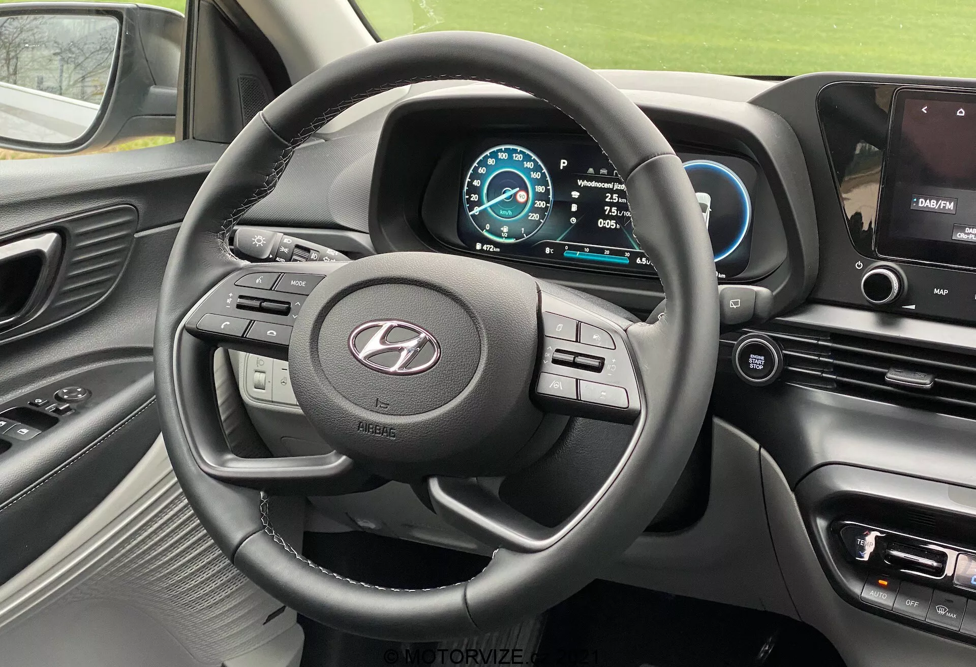 Vue intérieure de la Hyundai i20 2020 depuis le siège du conducteur, montrant le volant à trois branches avec plusieurs boutons de commande, le logo du fabricant bien en évidence, le tableau de bord numérique avec un schéma de couleurs bleu et blanc, le compteur de vitesse réglé sur 0 km/h, la sélection de vitesse en position Park, une partie de la console centrale avec écran d'infotainment affichant une carte de navigation, les boutons de commande pour le climat et les réglages du véhicule, un tableau de bord en matériau doux au toucher avec des accents métalliques autour des bouches d'aération et du volant, et une vue partielle de la bouche d'aération côté passager.