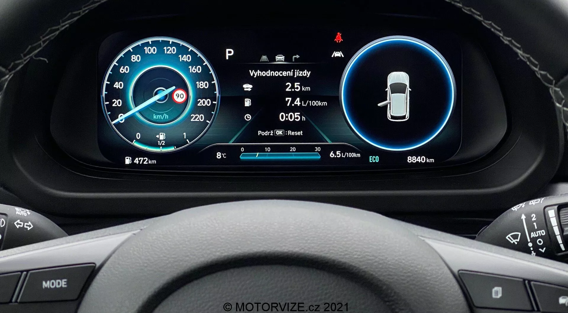 Perspective depuis le siège du conducteur de la Hyundai i20 2020 montrant une vue détaillée du tableau de bord numérique situé derrière le volant. Le design moderne comprend deux affichages circulaires principaux : un compteur de vitesse numérique et analogique à gauche et des indicateurs de fonctionnalités de sécurité à droite. Au centre, on trouve des informations supplémentaires telles que la température, l'odomètre, la consommation de carburant et la sélection de la vitesse. La périphérie affiche le kilométrage total (8840 km), la jauge de carburant et divers icônes systèmes. Le volant multifonction avec boutons de médias et de régulateur de vitesse est partiellement visible. En bas, un filigrane 'MOTORVIZE.cz 2021' est présent.
