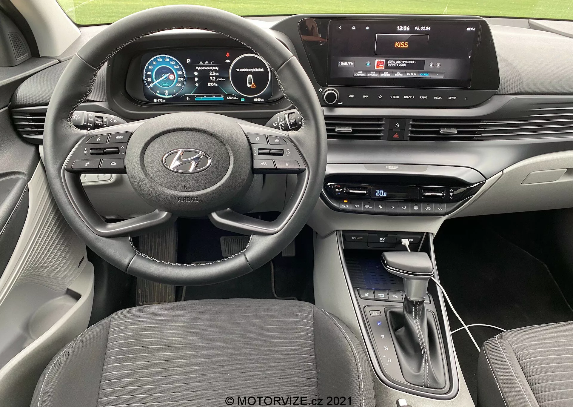 Vue intérieure de la Hyundai i20 2020 depuis la perspective du conducteur, présentant le volant à trois branches à fond plat avec des commandes intégrées pour l'audio, le régulateur de vitesse et la communication mains libres, un tableau de bord entièrement numérique avec des graphiques colorés et une console centrale équipée d'un système d'infodivertissement à écran tactile avec connectivité multimédia, des boutons et cadrans physiques pour le contrôle du climat, un levier de vitesse moderne et un design de tableau de bord contemporain en couches avec un schéma bicolore.