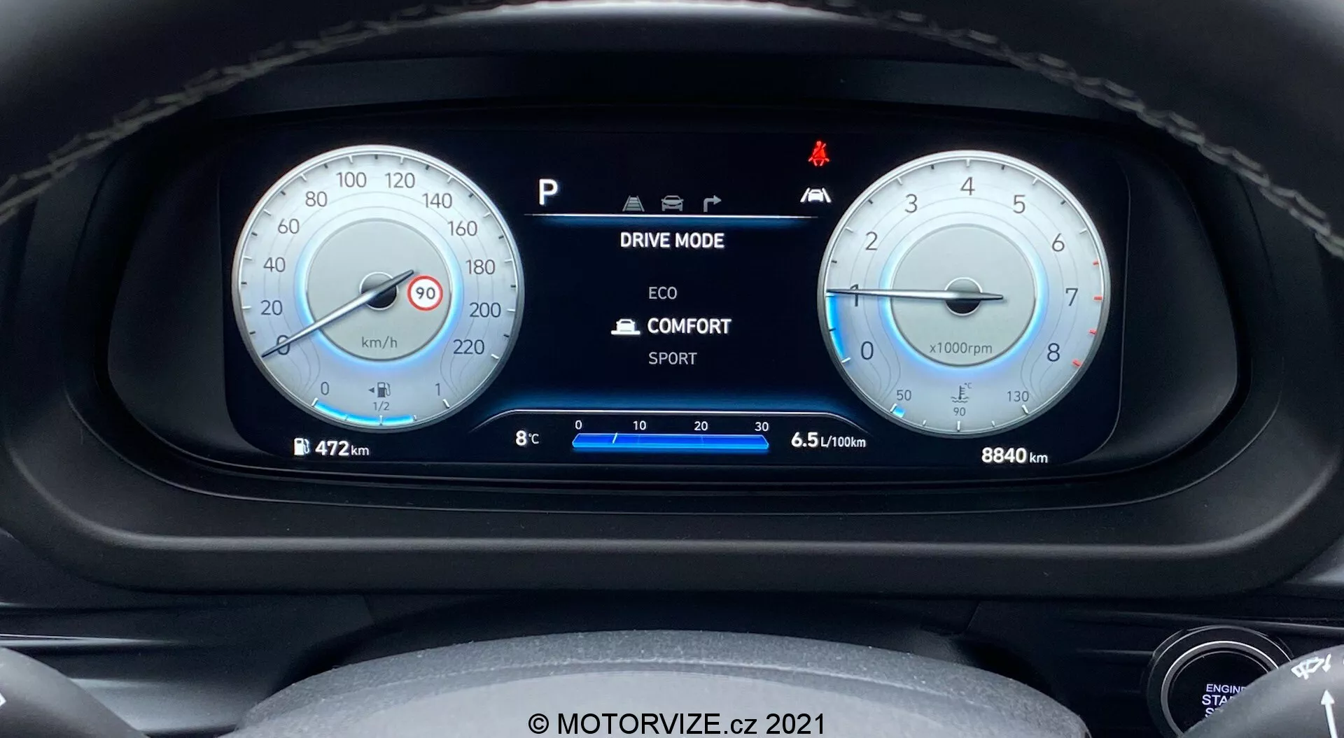 Vue rapprochée du tableau de bord de la Hyundai i20 2020 du point de vue du conducteur, avec deux jauges analogiques rondes principales et un affichage numérique central. La jauge de gauche affiche un compteur de vitesse marqué jusqu’à 260 km/h, tandis que celle de droite est un compte-tours avec une zone rouge à 6500 tr/min. L'affichage numérique central montre des options de mode de conduite, la température extérieure, la position du rapport et l'efficacité du carburant. L'odomètre indique 8840 km. Divers voyants lumineux sont visibles, y compris un pour la fonction de démarrage/arrêt automatique. L'image est nette et bien éclairée, avec un léger adoucissement autour des bords des jauges. Comprend le filigrane 'MOTORVIZE.cz 2021'.