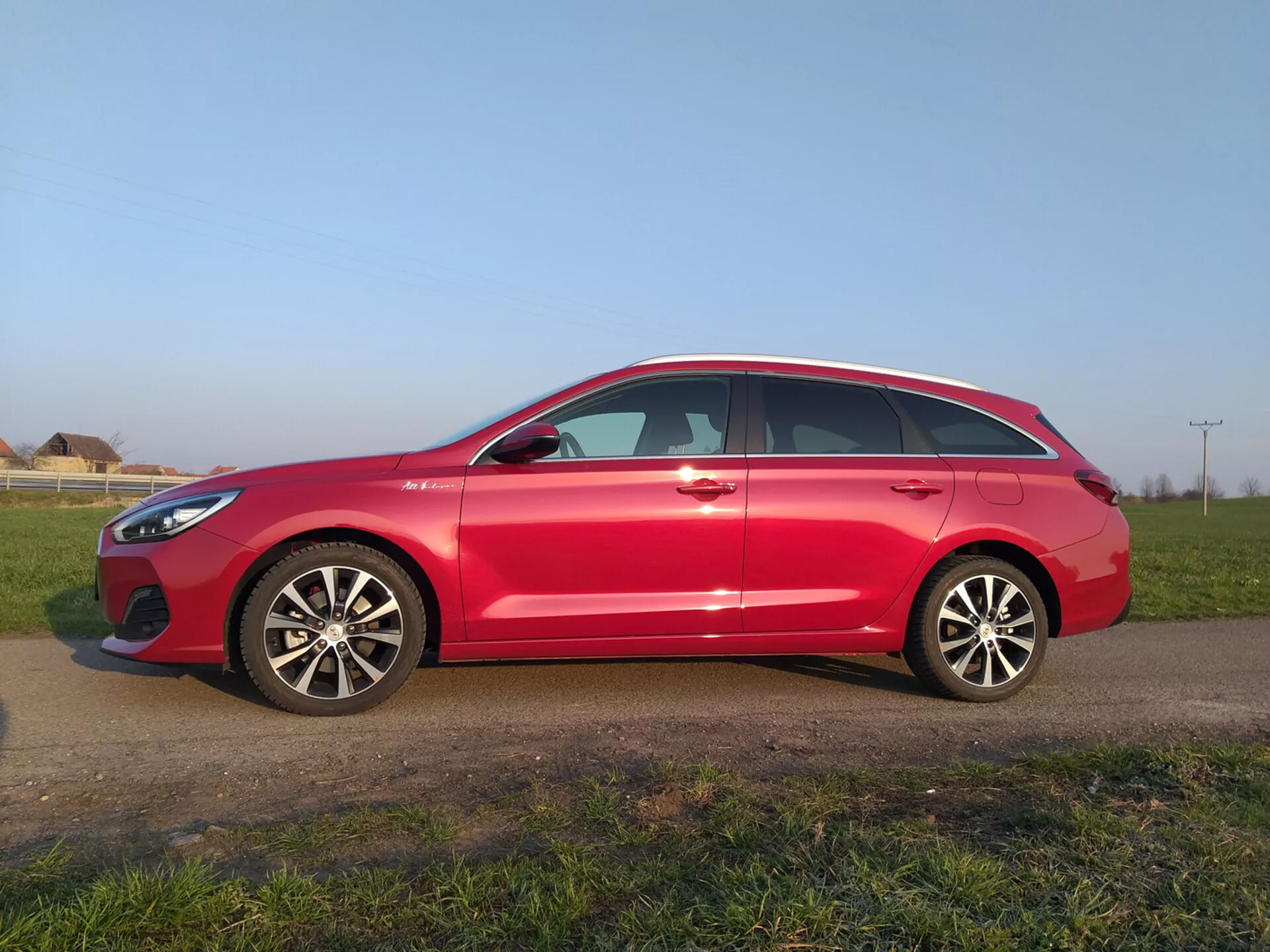 Hyundai i30 Fastback de 2017 en rouge, présentant un profil aérodynamique moderne avec une ligne de toit inclinée, jantes en alliage à multiples branches, rétroviseurs latéraux avec clignotants intégrés, fenêtres encadrées de chrome et pneus prêts pour la route, stationnée sur un champ herbeux sous un ciel dégagé.