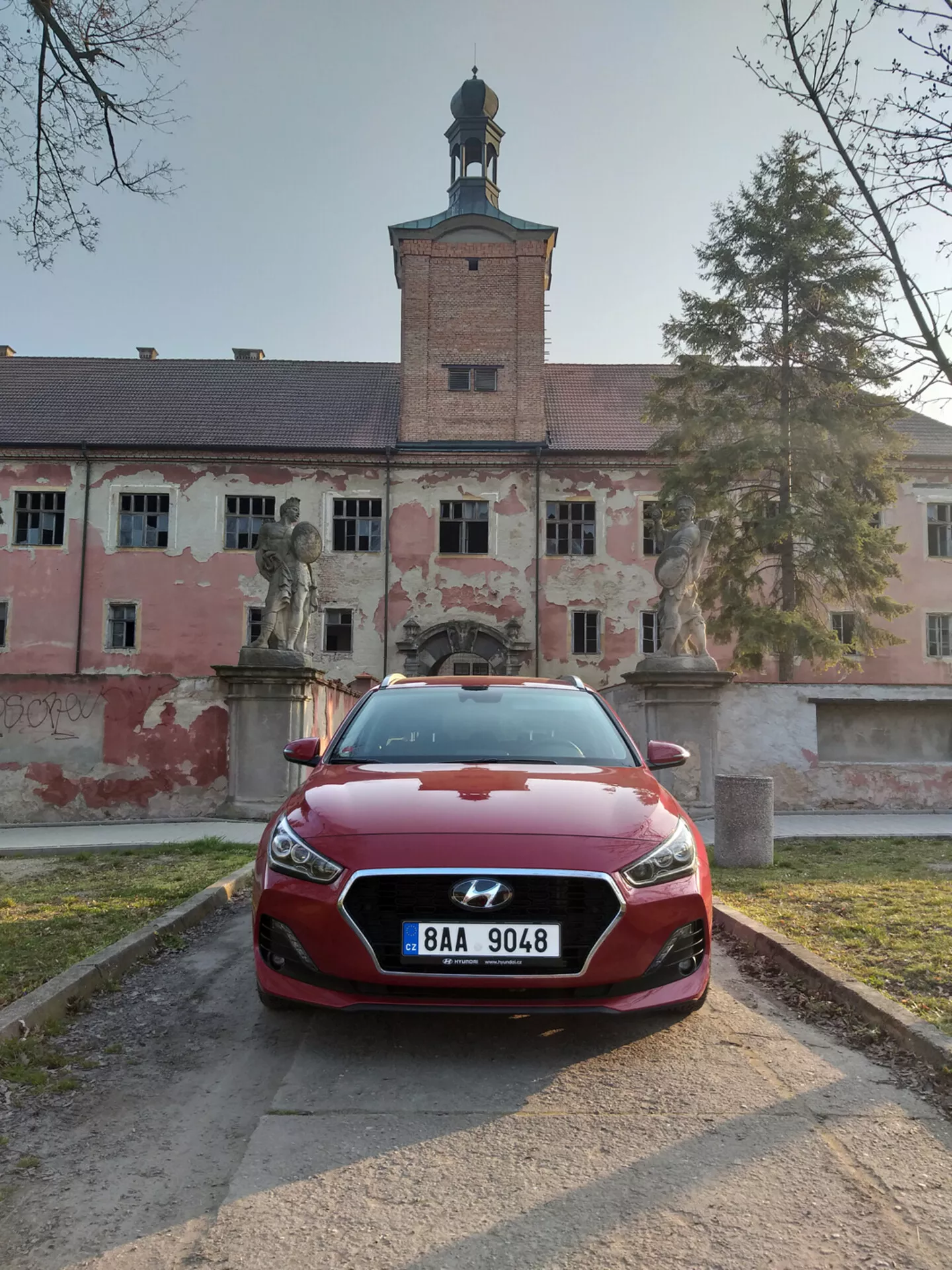 Hyundai i30 Fastback de 2017 en rouge, vue de face avec les phares LED et le design de la calandre sculpté, positionnée sur un chemin de gravier devant un bâtiment historique.