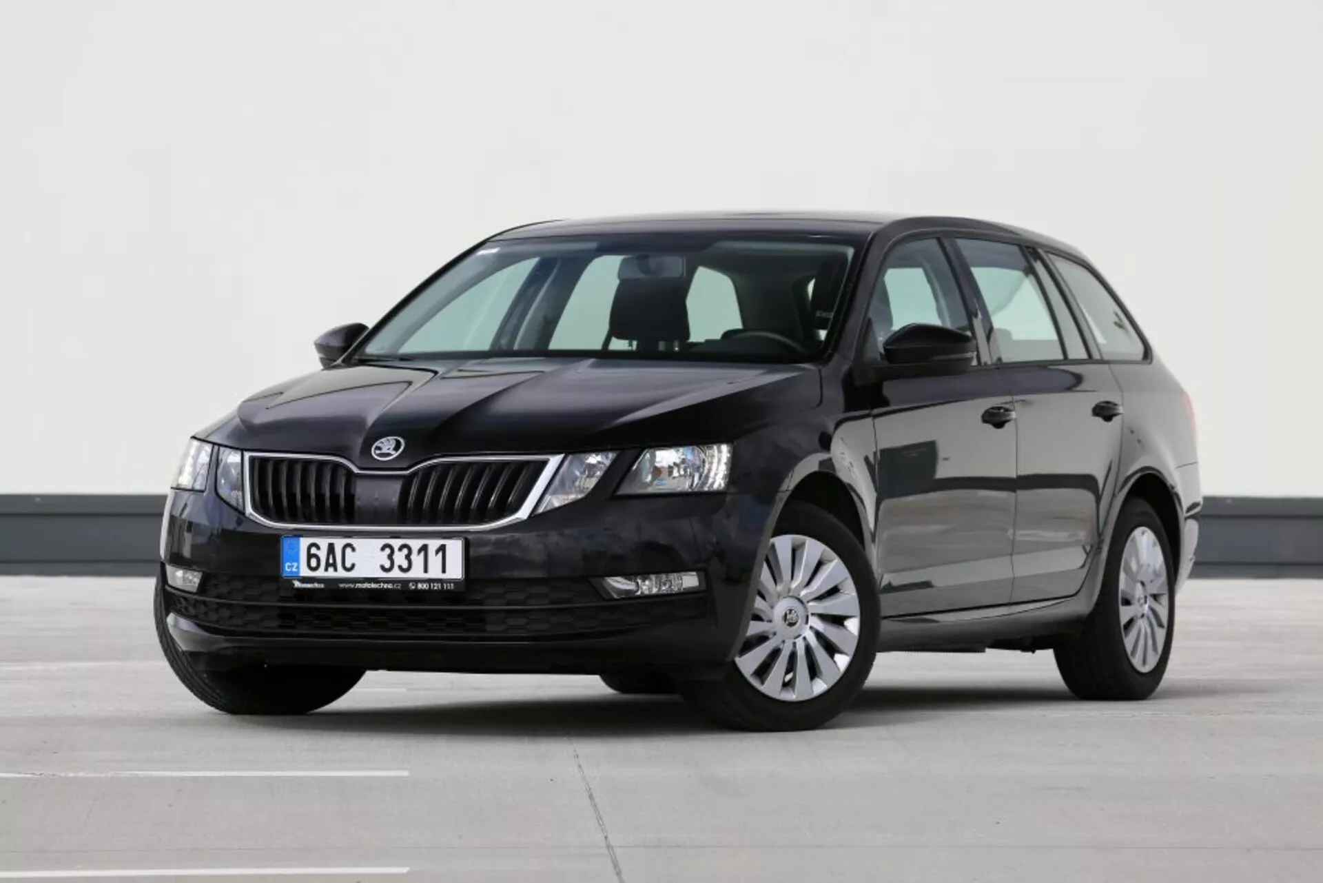 Véhicule Škoda stationnaire vu depuis une perspective frontale des trois quarts côté conducteur, affichant la grille, les phares, le capot, le pare-brise, les portes du conducteur et du passager arrière, une partie du toit et le miroir latéral gauche sur une surface plane intérieure avec éclairage professionnel