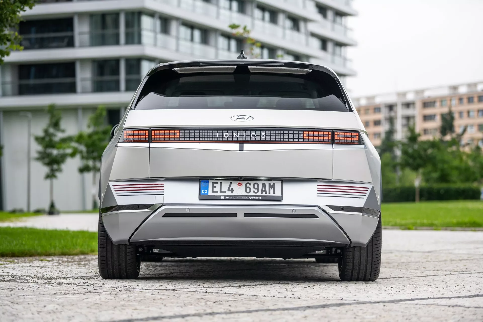 Vue arrière de la Hyundai Ioniq 5 2WD 2021, mettant en évidence une barre lumineuse arrière distinctive sur toute la largeur, un logo du fabricant centré, un arrière plat avec des lignes horizontales nettes, un pare-chocs arrière minimaliste avec des bandes réfléchissantes et une plaque d'immatriculation au format européen. Le véhicule est centré sur une surface asphaltée avec du feuillage et des bâtiments modernes en arrière-plan, sous une lumière nuageuse.