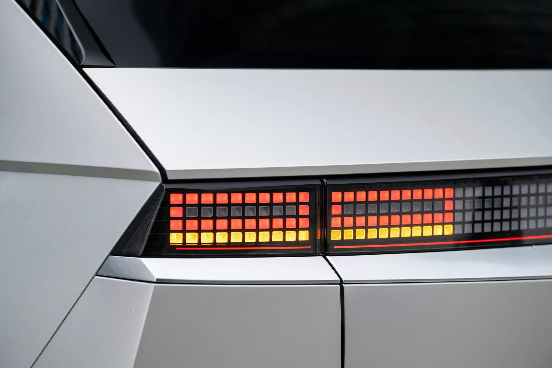 Image de gros plan de l'assemblage des feux arrière de la Hyundai Ioniq-5 2021 2WD. Focalisation sur les lumières LED rouges et pixelisées dans une vue rapprochée, mettant en valeur la carrosserie géométrique de la voiture en blanc ou argent clair. Vue détaillée des éléments de design modernes et de la technologie d'éclairage.
