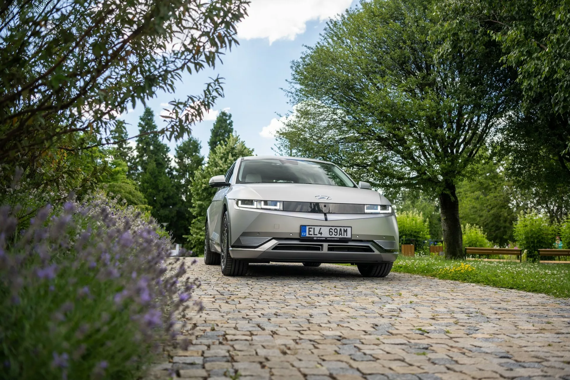 Vue en contre-plongée d'une Hyundai Ioniq 5 2WD 2021 garée à l'extérieur sur une surface pavée. La caméra se concentre sur l'avant de la voiture, y compris la calandre, les phares avant gauche et une partie du capot et du pare-brise, avec un arrière-plan d’arbres verts, de buissons et de fleurs violettes sous une lumière naturelle abondante.