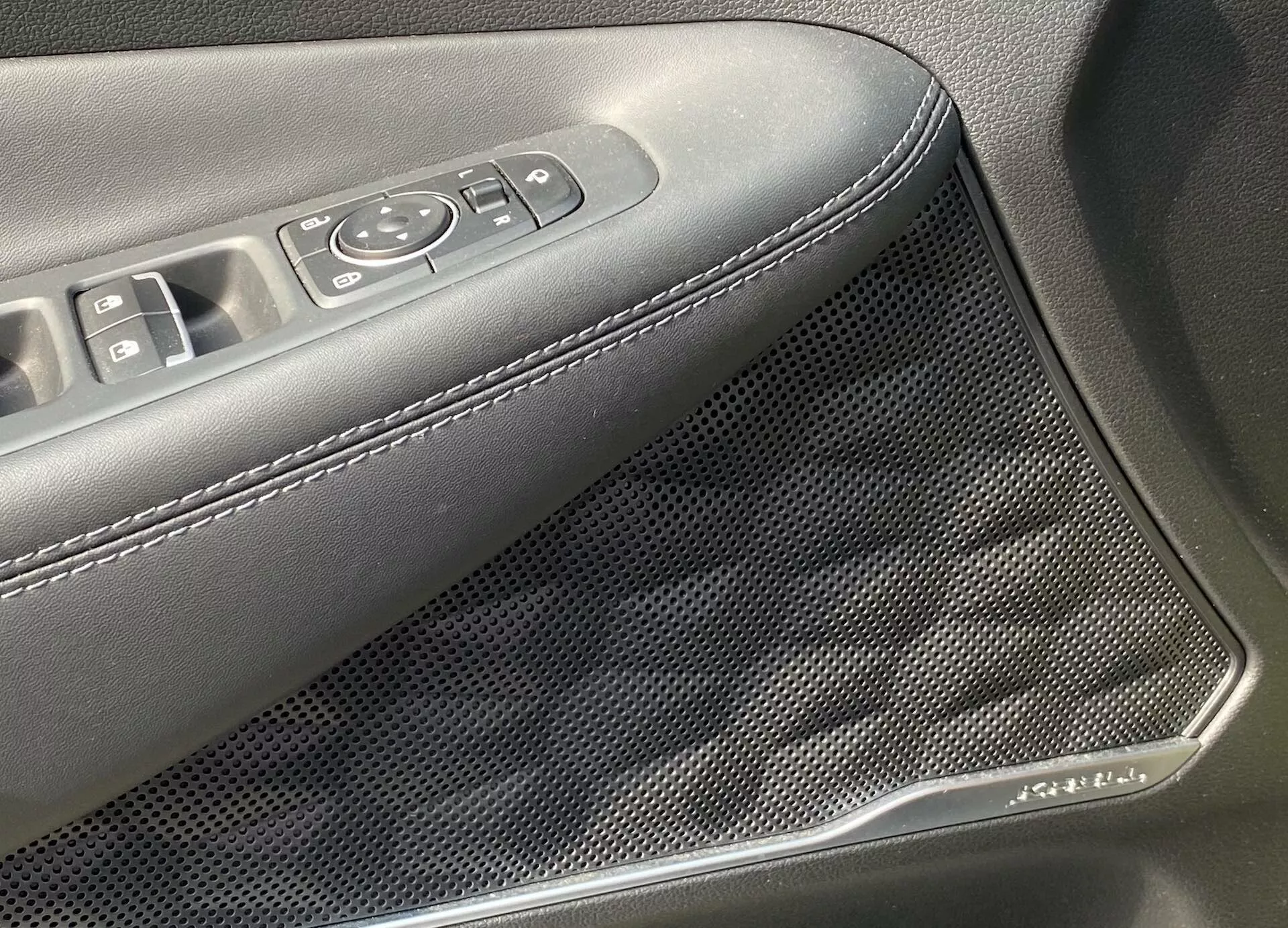 Vue en gros plan du panneau de porte intérieur d'une Hyundai Santa Fe 2020, mettant en lumière la zone avec les commandes des vitres électriques et des rétroviseurs, ainsi qu'une grille de haut-parleur à motif nid d'abeille. L'accoudoir est en matériau foncé avec des coutures blanches. Une garniture chromée orne la grille du haut-parleur, révélant des caractéristiques avancées de l'audio et du design esthétique.