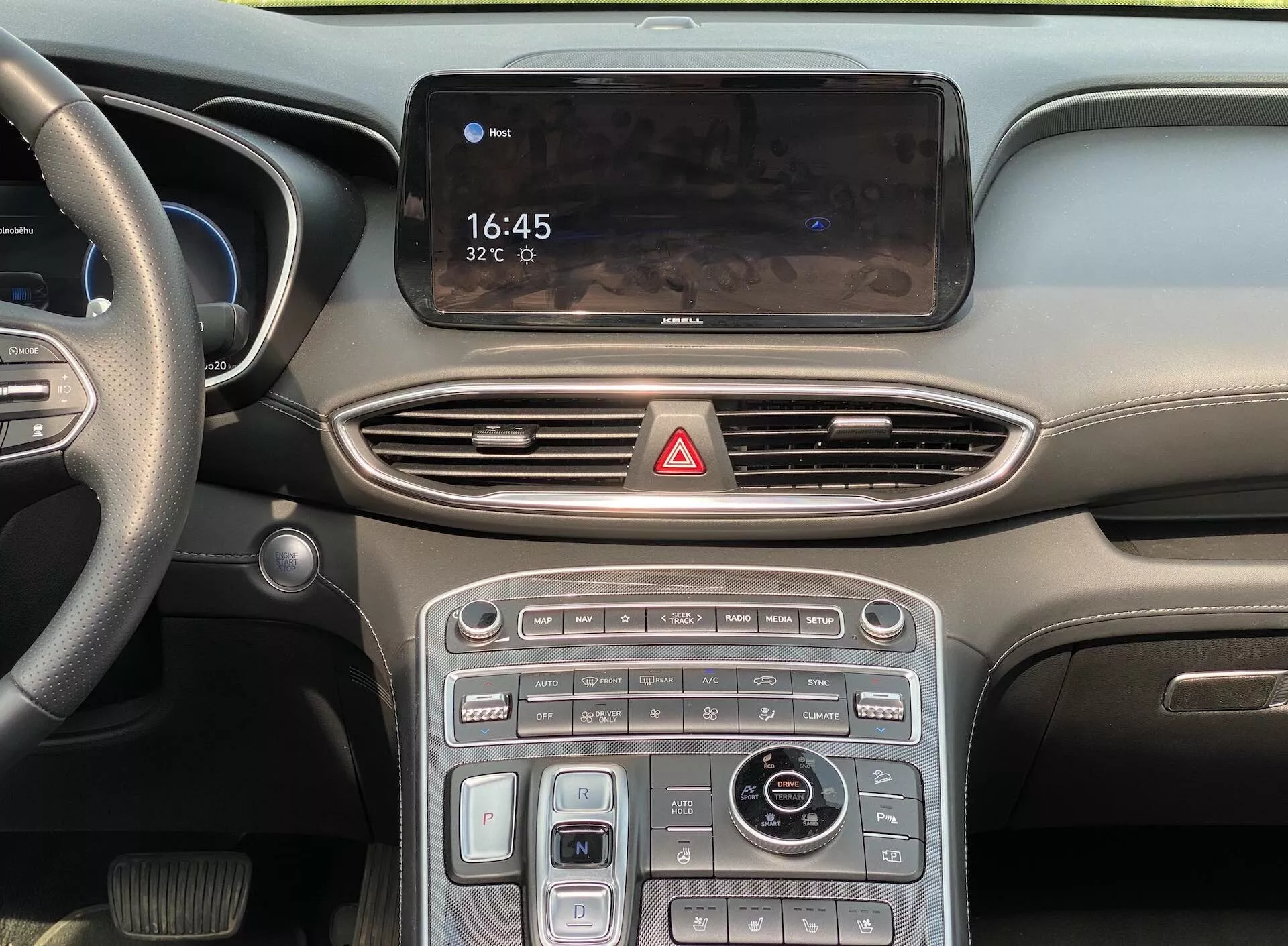 Vue rapprochée et en gros plan de la console centrale et d'une partie du tableau de bord d'une voiture moderne, incluant l'affichage de l'infotainment montrant 16:45 et 32°C, des bouches d'aération fluides, un bouton de feux de détresse, un mélange de commandes tactiles et de boutons physiques avec des accents métalliques, un cadran rotatif pour le contrôle de l'infotainment et de la navigation, et un sélecteur de mode de conduite, le tout suggérant un intérieur élégant et moderne avec des fonctionnalités avancées telles que le contrôle de la climatisation, le chauffage/refroidissement des sièges et plusieurs modes de conduite. Le tableau de bord flou et la vue partielle du volant à gauche impliquent un véhicule haut de gamme conçu pour les marchés avec conduite à gauche.