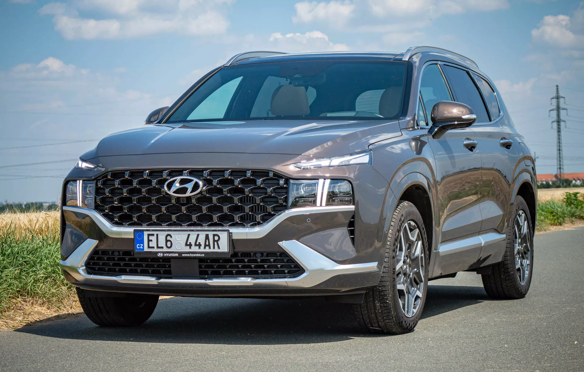 L'image montre un SUV Hyundai Santa Fe 2020 garé sur une route en asphalte, capturé depuis un angle de trois quarts avant gauche. Elle met en évidence la grande calandre avec un motif spécifique à la marque et un logo central, les phares LED élégants et le capot sculpté. Le pare-brise est large et offre une grande visibilité, et le profil latéral révèle des jantes en alliage à plusieurs branches et le rétroviseur côté conducteur avec des clignotants intégrés potentiellement. Le véhicule de couleur foncée dégage une apparence sophistiquée avec ses éléments de design modernes, y compris éventuellement des capteurs ou des caméras avant.