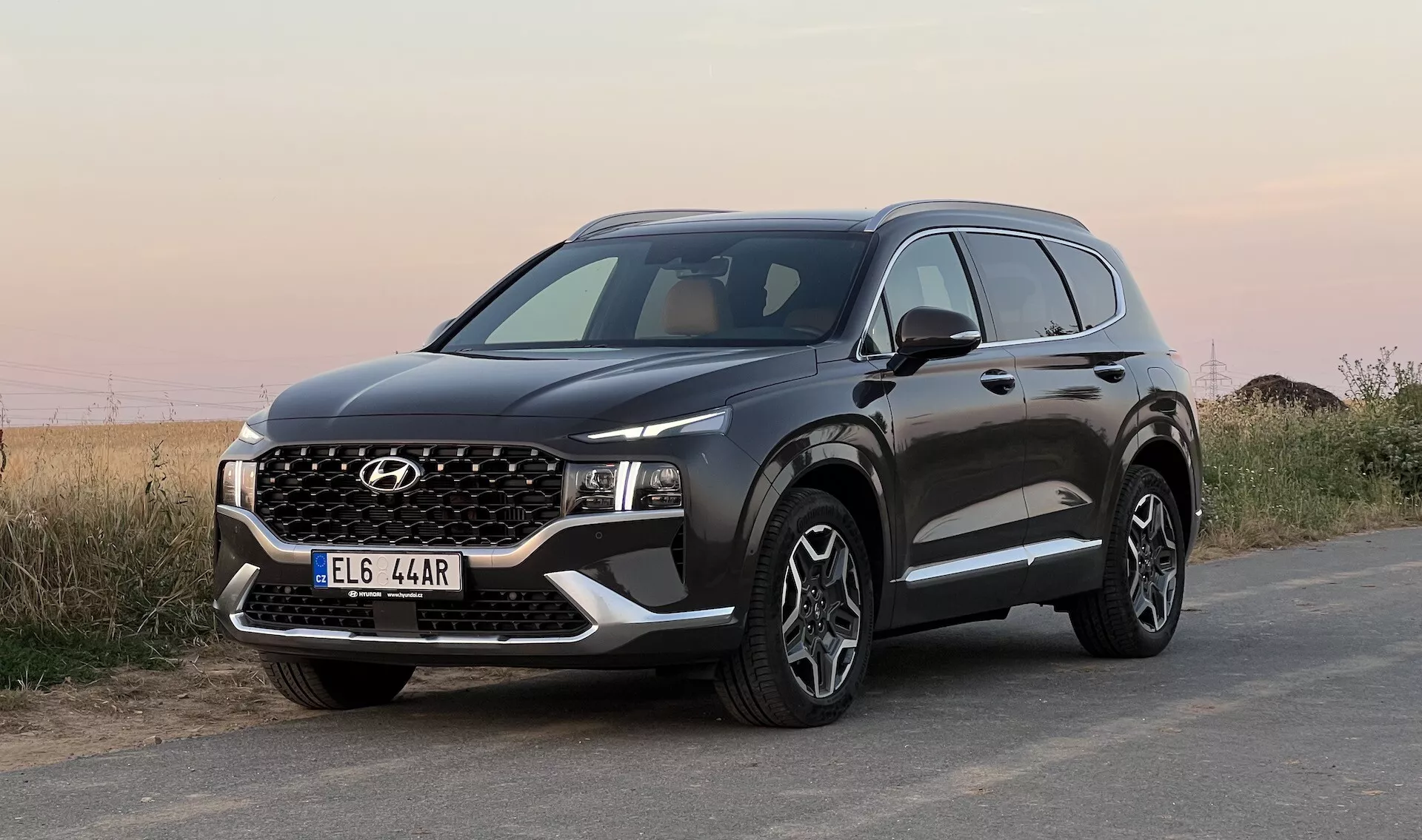 Image d'un SUV Hyundai Santa Fe 2020 stationné sur une surface asphaltée, prise sous un angle bas avec une vue frontale trois quarts, affichant la calandre avant, des phares LED étroits, un pare-choc avec anti-brouillards et une fausse plaque de protection, un capot avec des plis angulaires, de grandes roues en alliage multibranches, des protections noires sur les arches des roues et le bas des portes, un rétroviseur latéral avec clignotants intégrés et des fenêtres encadrées de chrome. L'arrière-plan présente un paysage naturel indéfini sous un ciel ouvert.